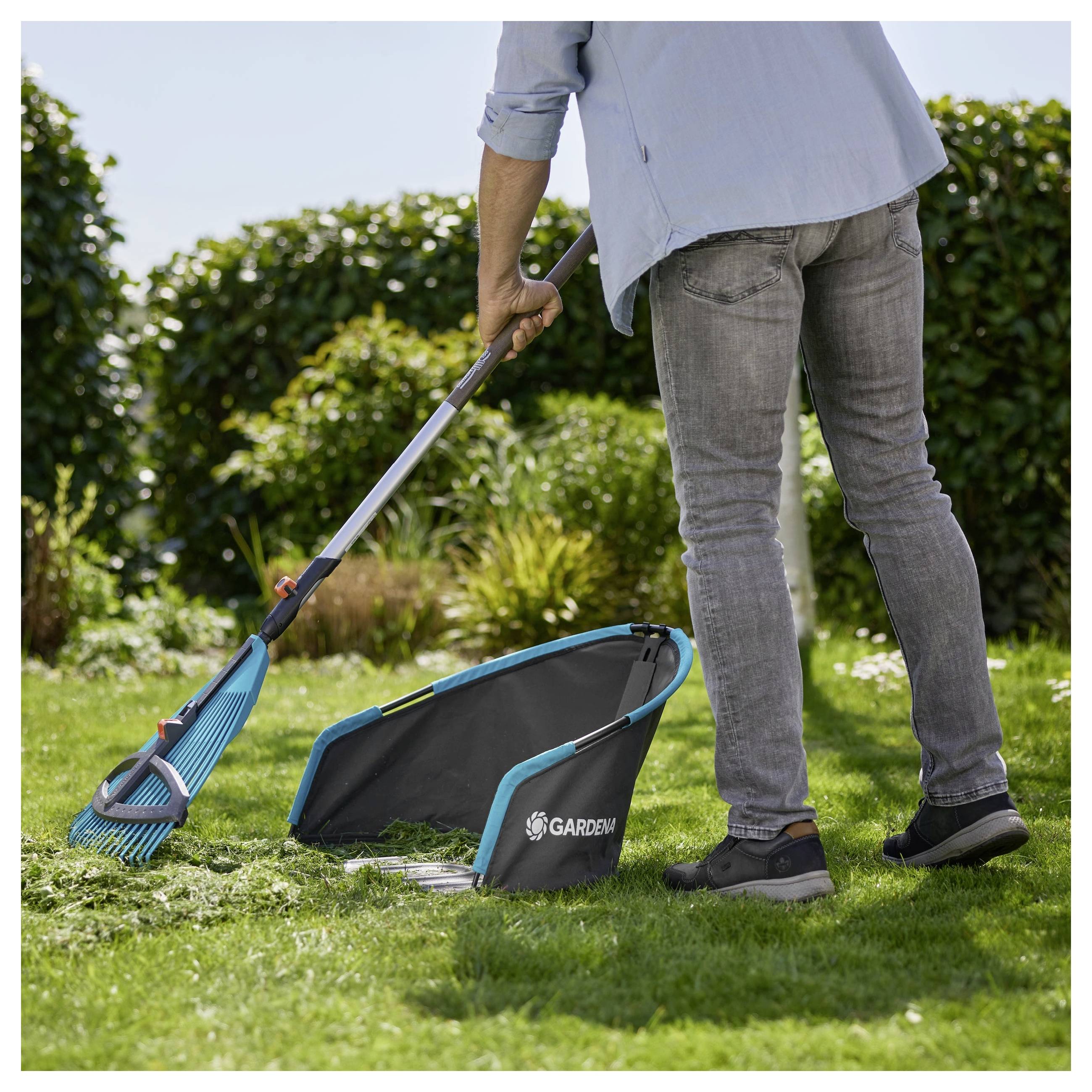 Eine Person harkt Gras in einen Sammelbeutel mit der Aufschrift "Gardena" in einer Gartenumgebung, umgeben von grünen Sträuchern und klarem blauem Himmel.