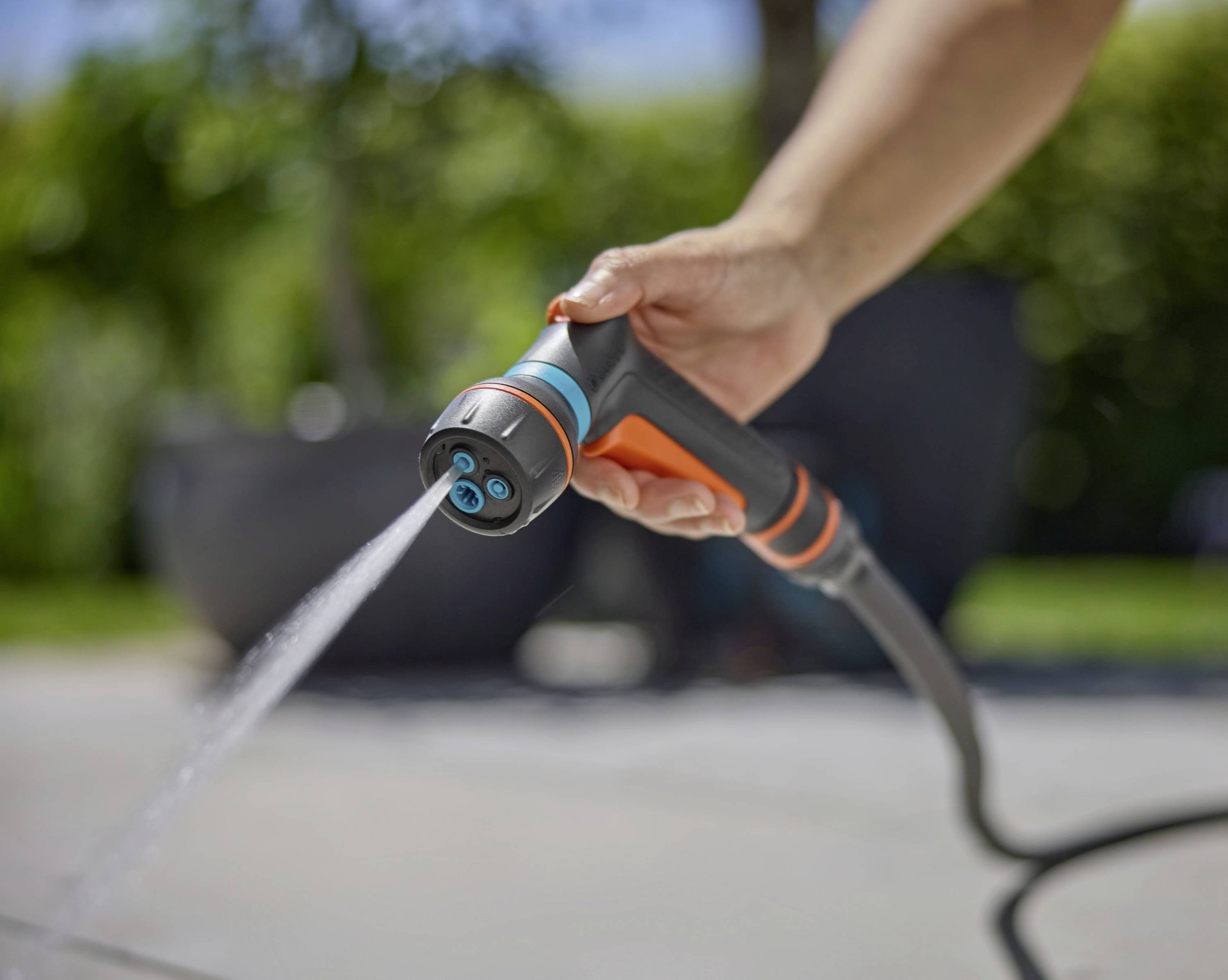 Eine Hand, die eine Gartenschlauch-Sprühbrause hält und einen Wasserstrahl freisetzt, vor einem unscharf verschwommenen grünen Hintergrund.