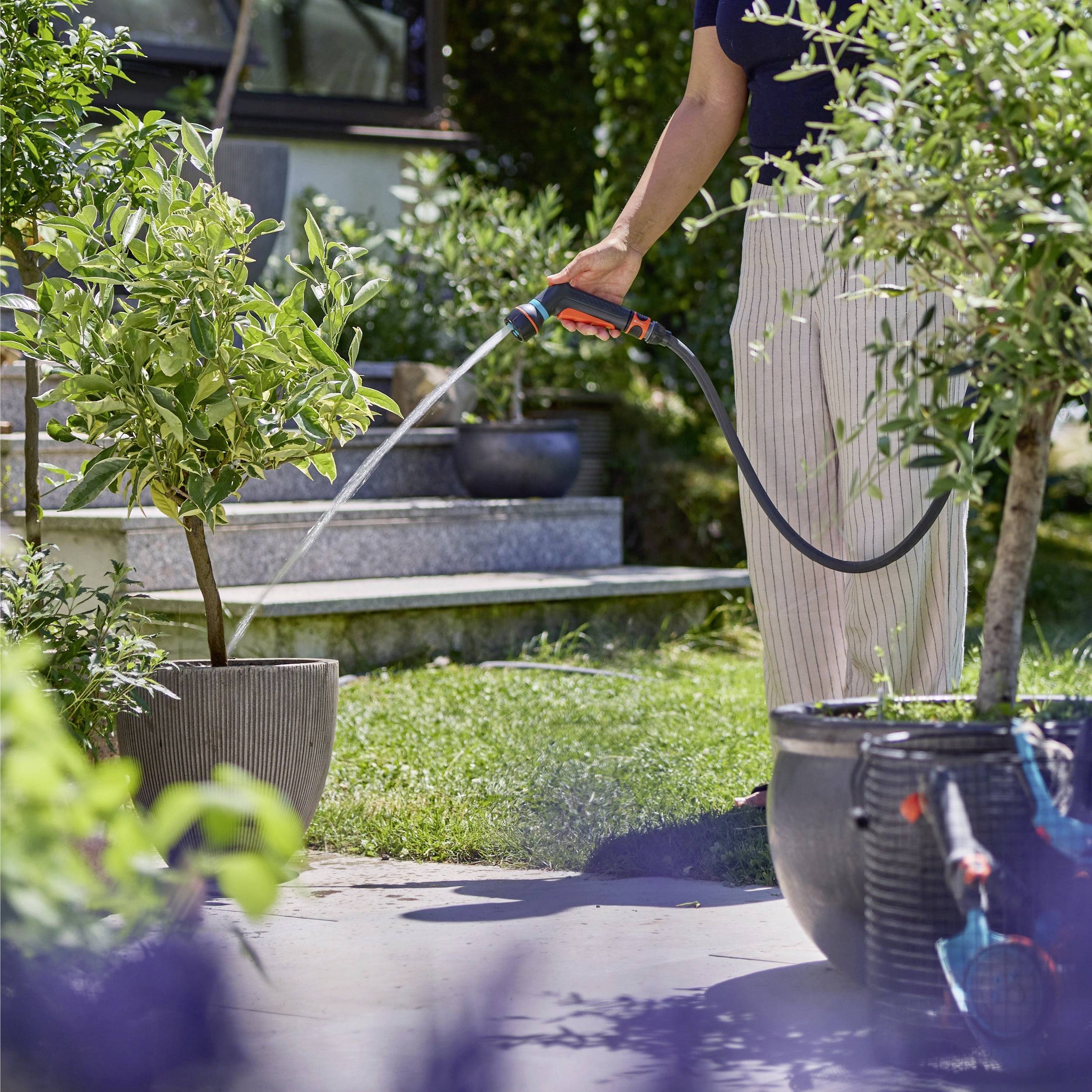 Eine Nahaufnahme einer Hand, die eine Gartenschlauchdüse hält und Wasser auf eine Terrasse spritzt, mit verschwommener Vegetation im Hintergrund.