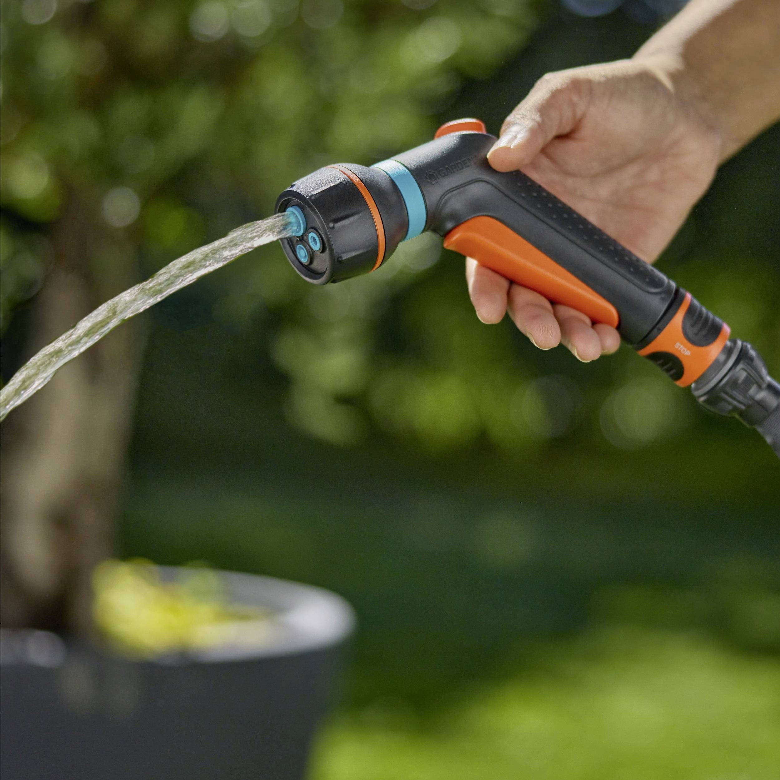 Eine Hand hält eine orange und blaue Gartenschlauchbrause, die Wasser versprüht, während im unscharfen Hintergrund eine Topfpflanze zu sehen ist.