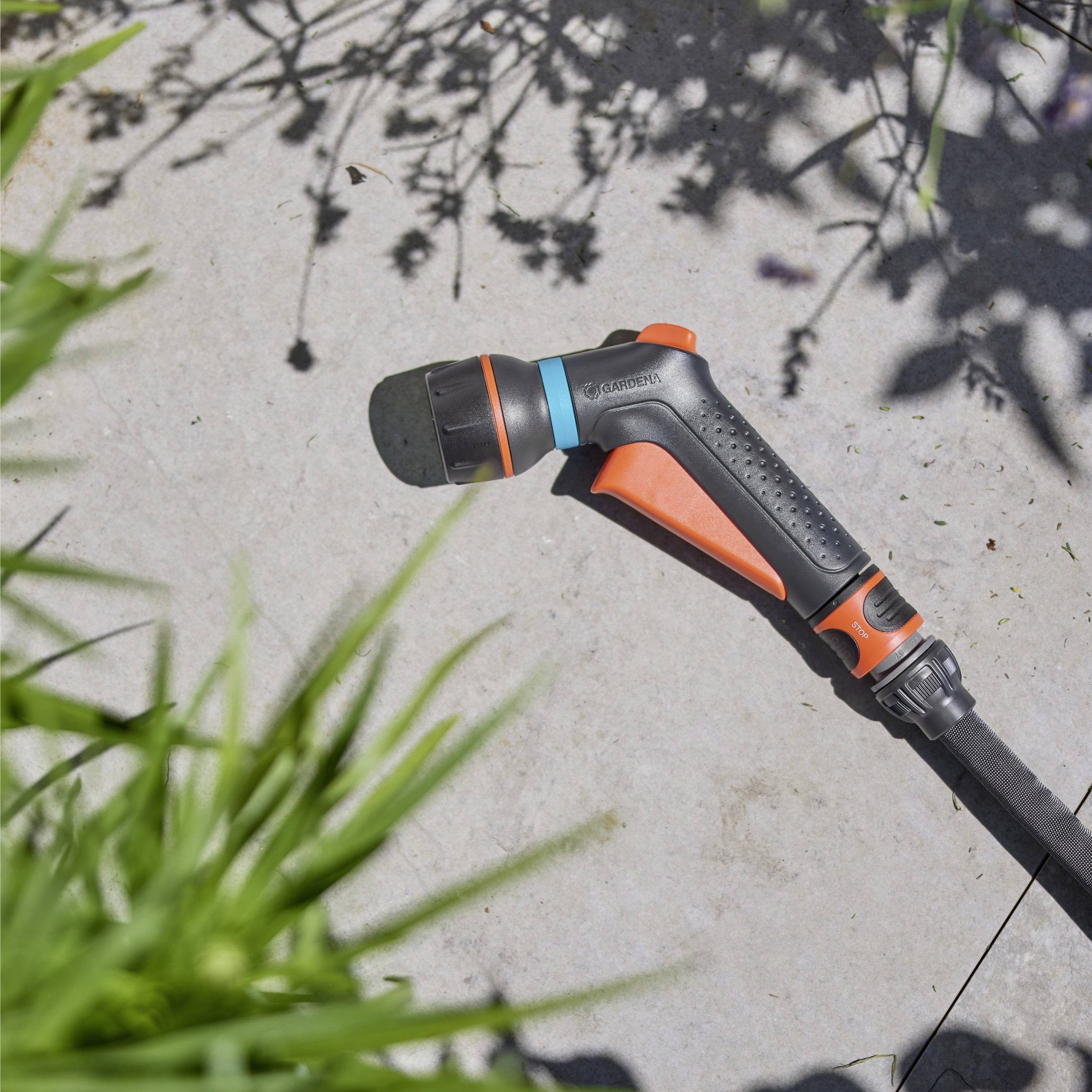 Eine Gartenschlauch-Sprühbrause mit ergonomischem Griff liegt auf einer Steinterrasse, umgeben von Gras und Pflanzenschatten, was auf Gartenbewässerung hindeutet.