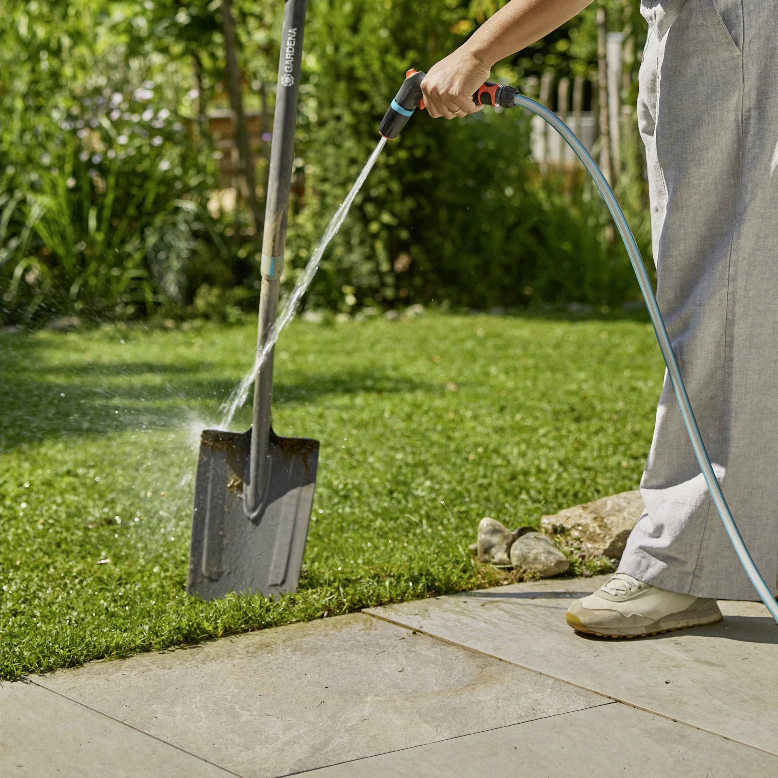 Eine Person bewässert Gras mit einem Gartenschlauch, neben einer Schaufel, an einem sonnigen Tag in einem Garten, was auf eine Gartenarbeit oder Rasenpflegeaktivität hindeutet.
