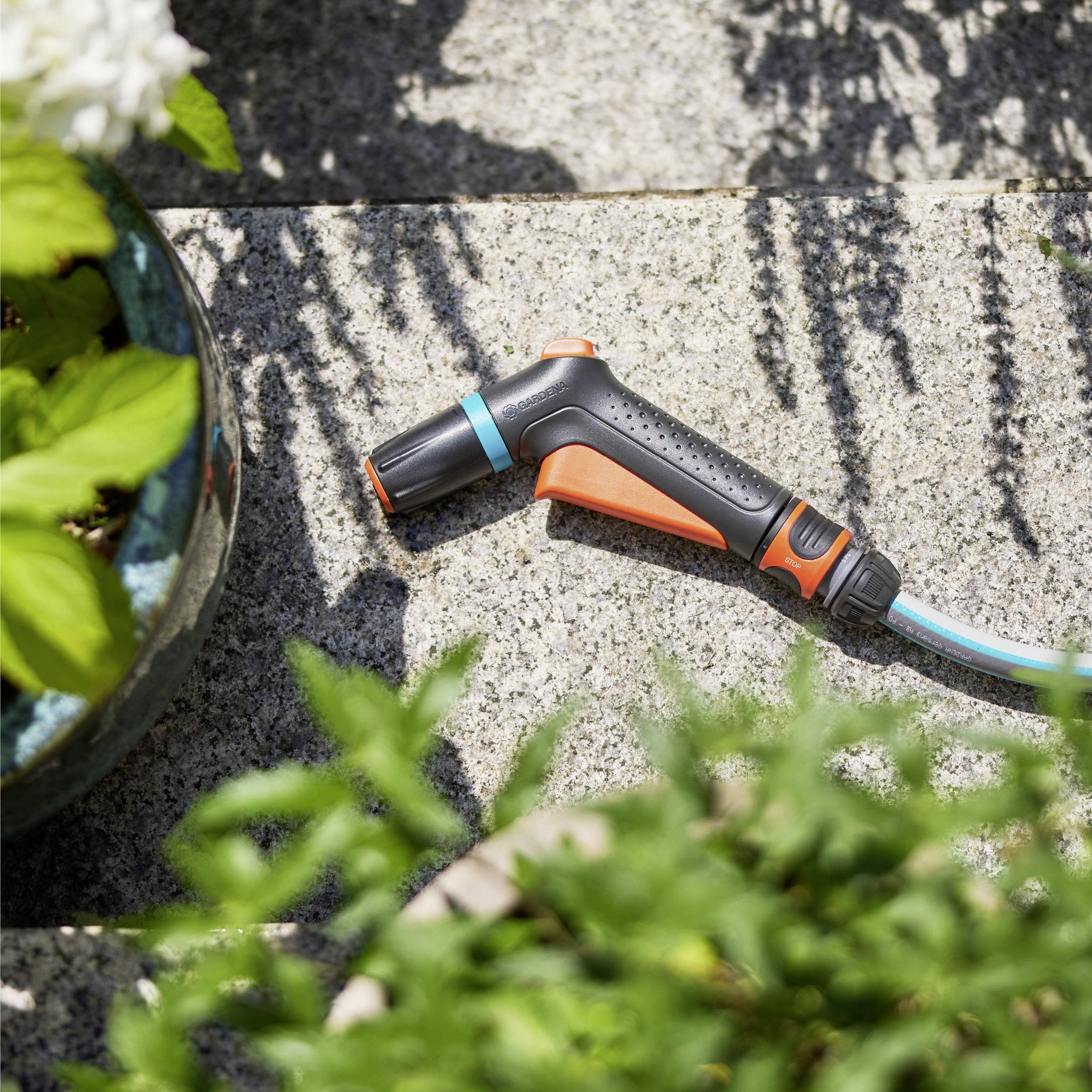 Eine Gartenschlauch-Düse mit orange-schwarzem Design liegt auf einer Steinoberfläche neben Topfpflanzen und ist bereit zum Bewässern.