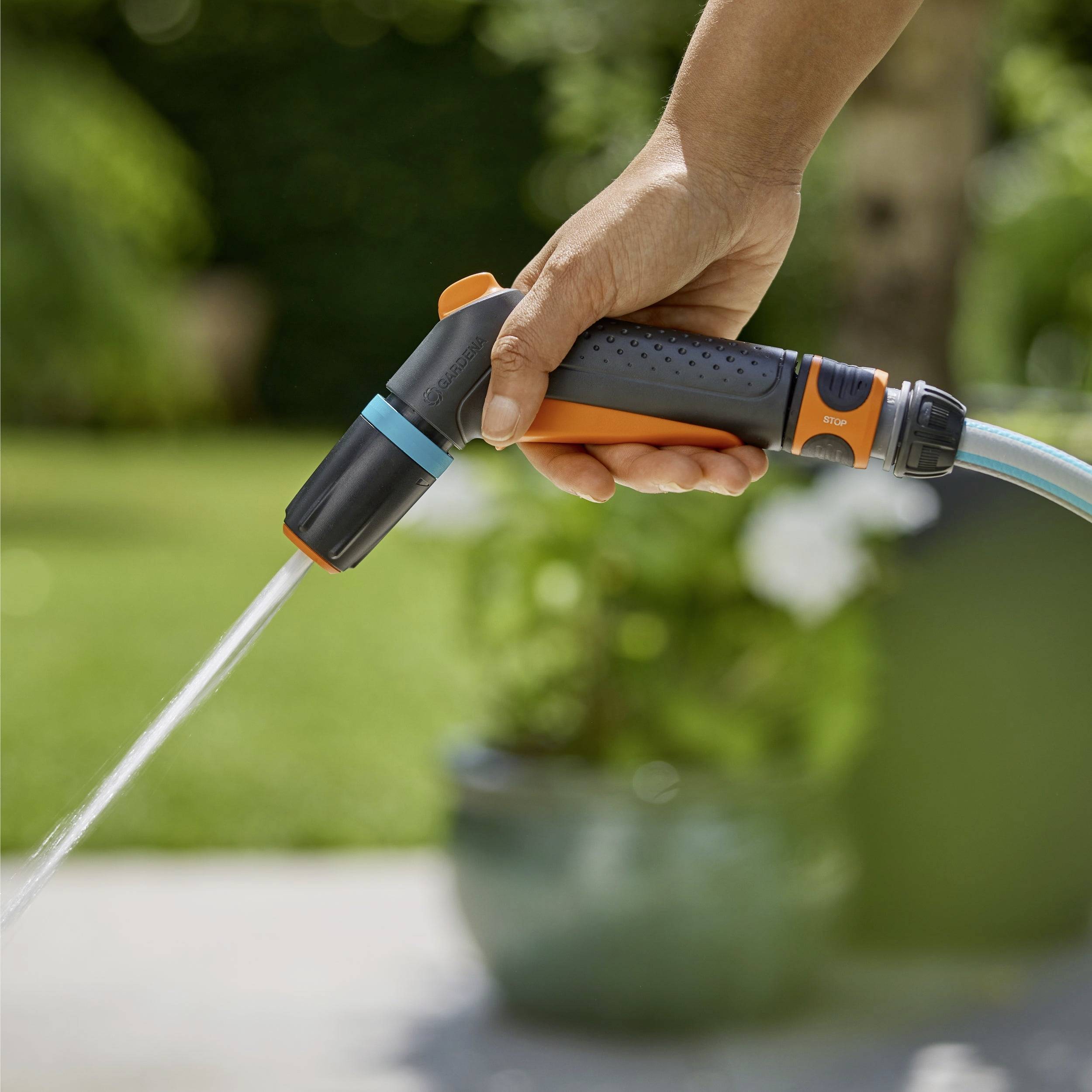Eine Hand hält einen Gartenschlauch mit einer Düse und richtet einen Wasserstrahl auf einen verschwommenen grünen Gartenhintergrund.