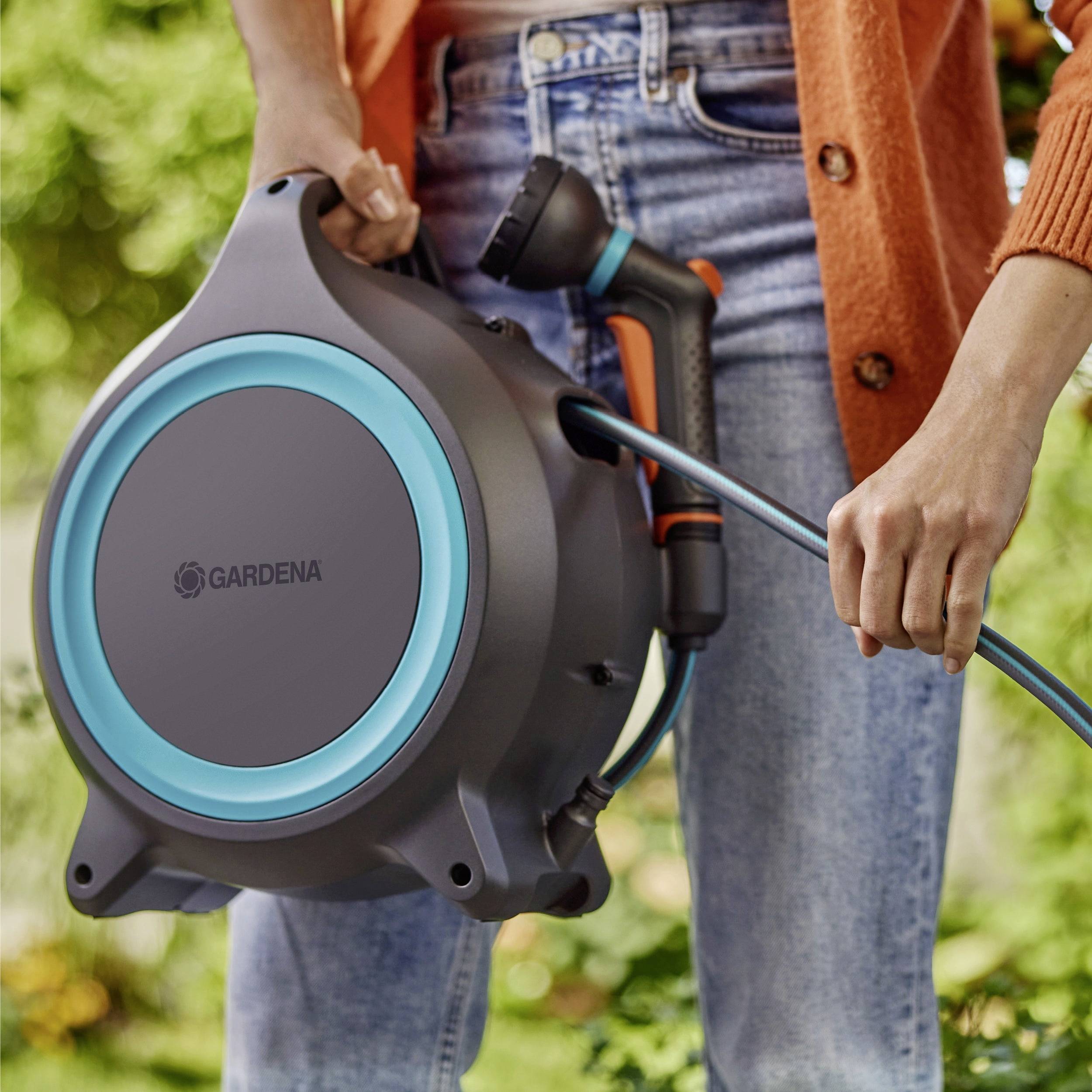 Person, der eine graue und blaue GARDENA-Schlauchtrommel in einem Garten hält und einen orangefarbenen Pullover und Jeans trägt.