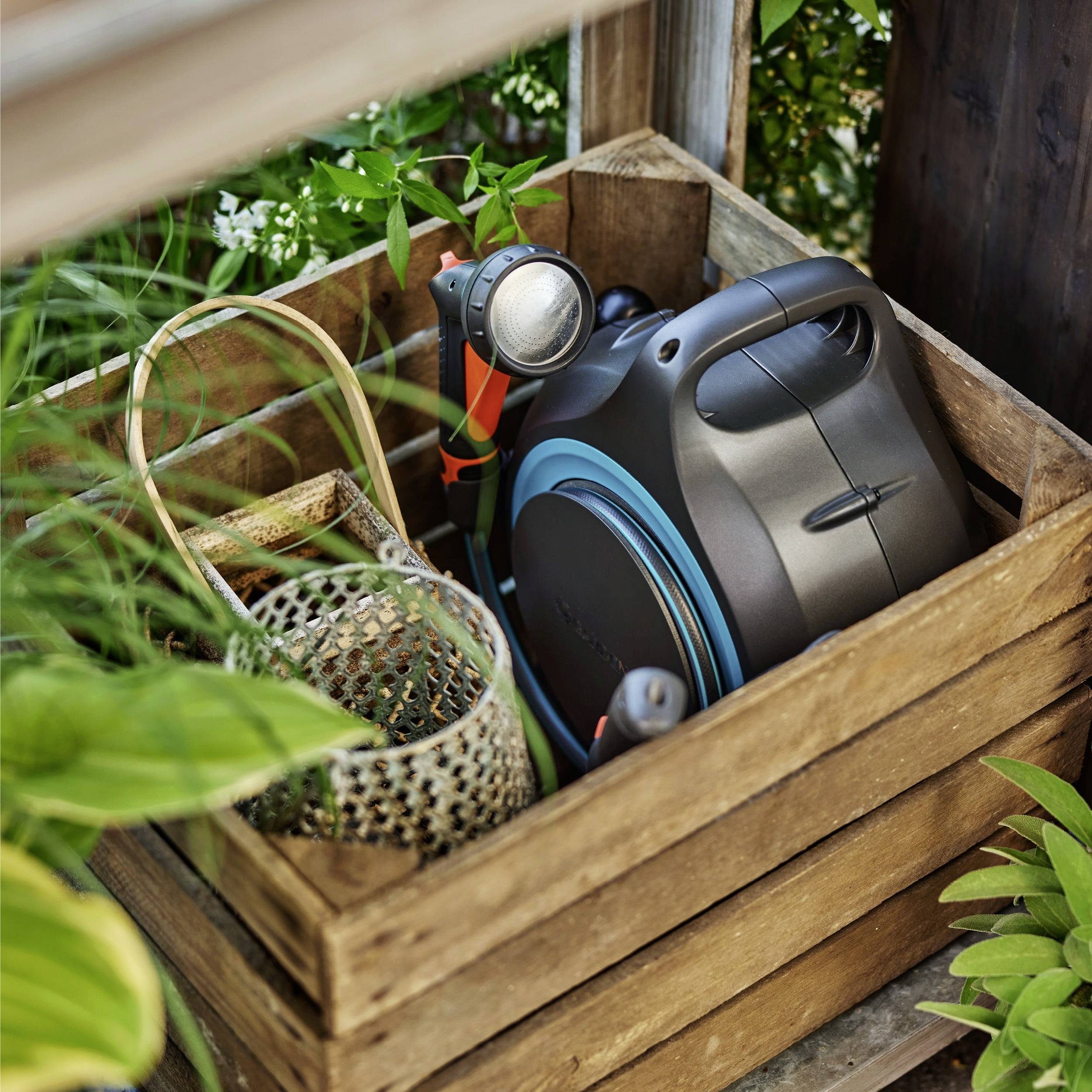 Ein Gartenschlauch-Wagen und eine Düse sind ordentlich in einer Holzkiste verstaut, umgeben von üppigem grünem Laub in einer Gartenumgebung.