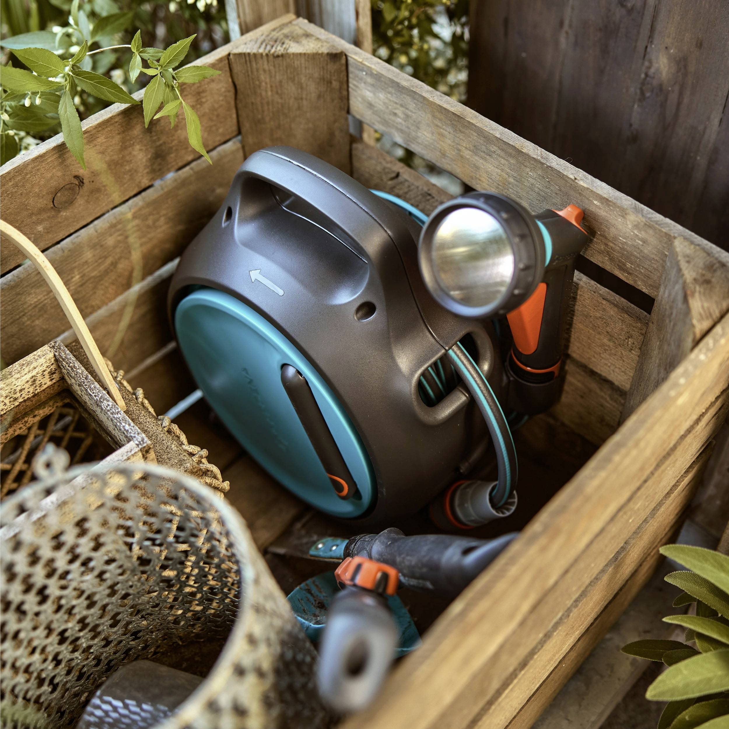 Gartenschlauchrolle mit angebrachter Sprühvorrichtung, gelagert in einer Holzkiste, umgeben von Gartengeräten und Pflanzen, was eine Gartenarbeitsumgebung andeutet.
