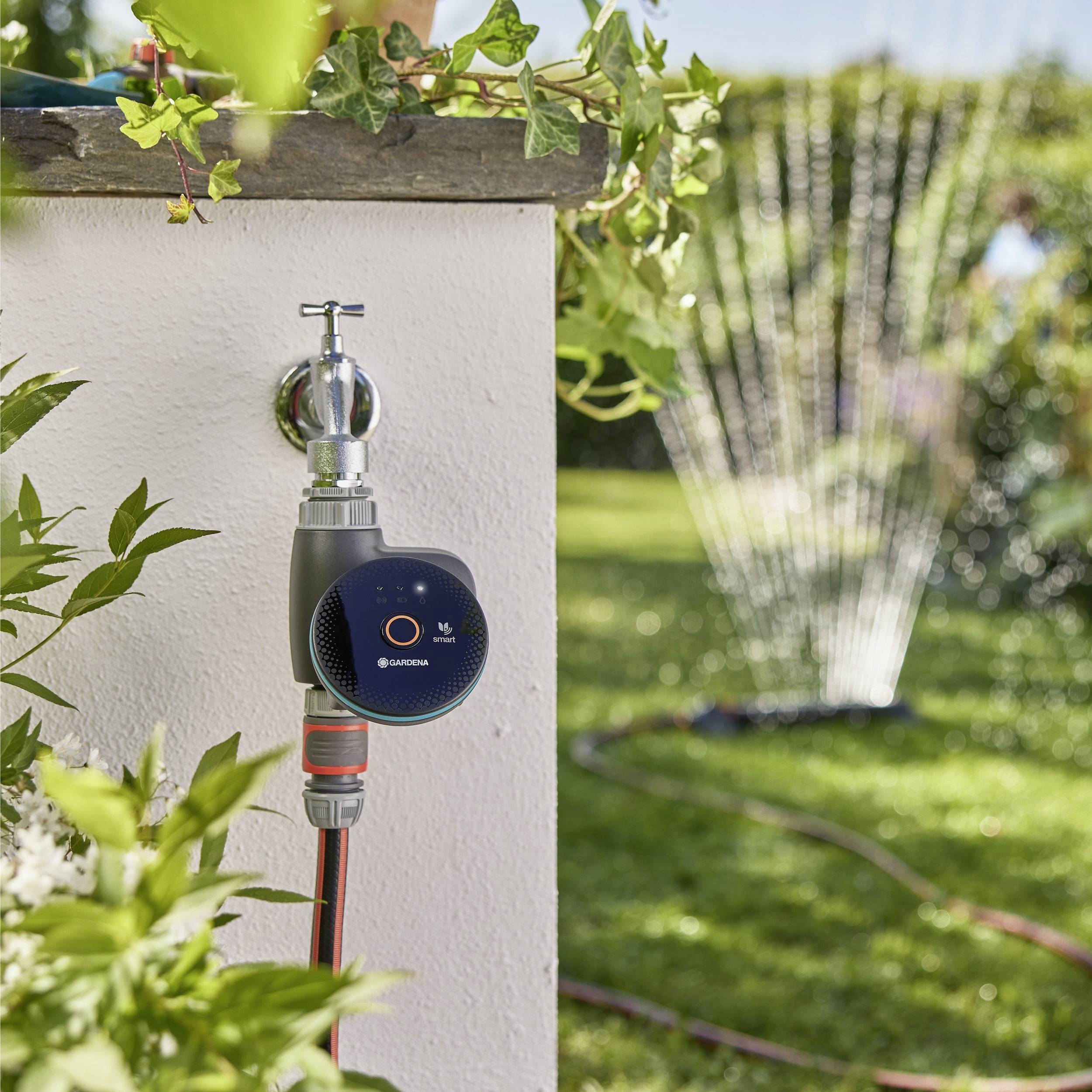 Ein intelligenter Wassertimer, der an einem Außenwasserhahn befestigt ist, steuert einen Gartenschlauch und bewässert an einem sonnigen Tag den Rasen im Hintergrund.