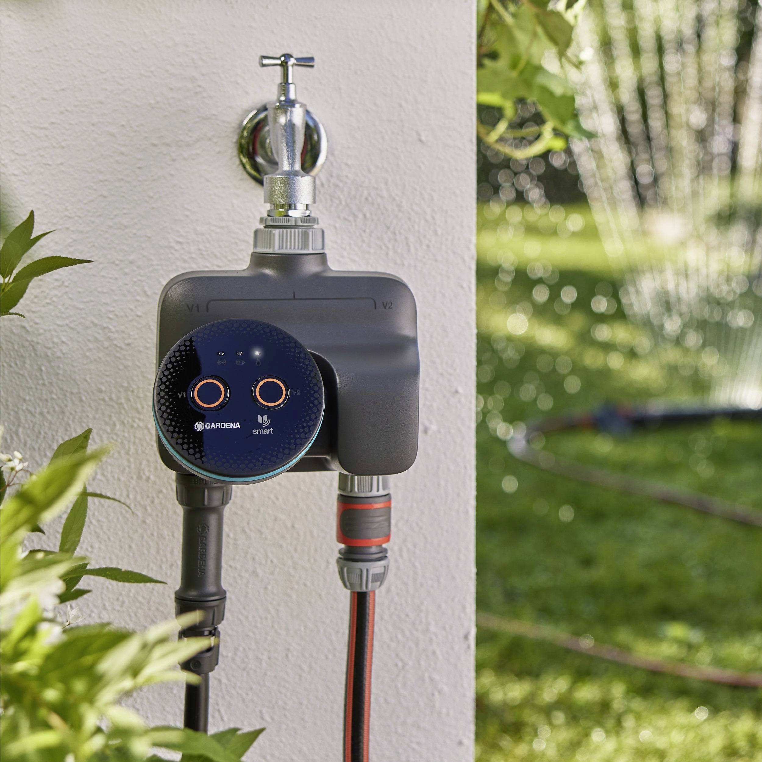 Automatischer Wasserzeitschalter, der an einem Außenwasserhahn befestigt ist und Gartenbewässerungssprinkler reguliert. Grüne Blätter und Wassersprühnebel sind im Hintergrund sichtbar.