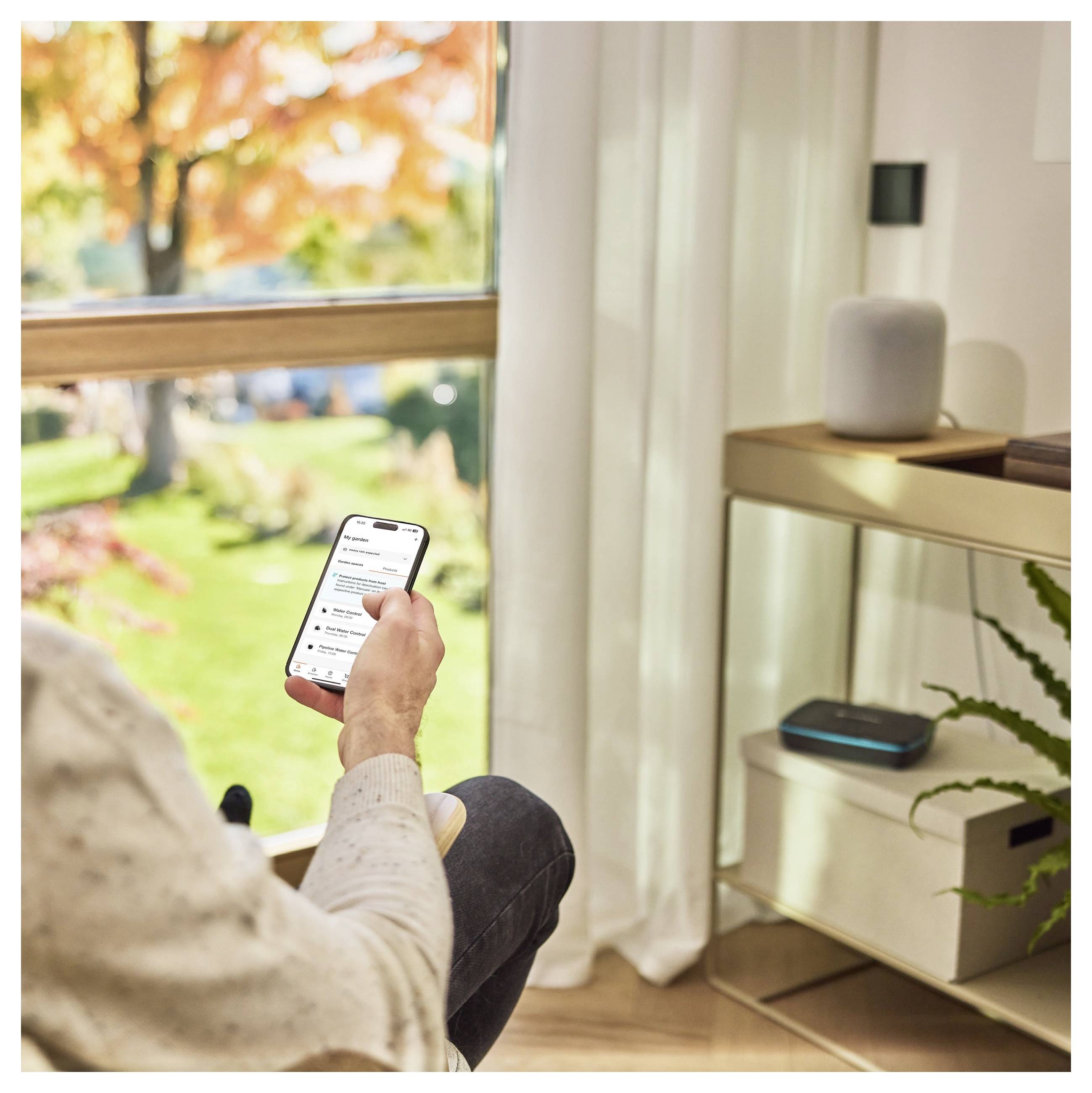 Eine Person sitzt am Fenster und hält ein Smartphone mit einer Liste. In der Nähe stehen Smart-Lautsprecher auf einem Regal. Draußen ist leuchtend buntes Herbstlaub zu sehen.