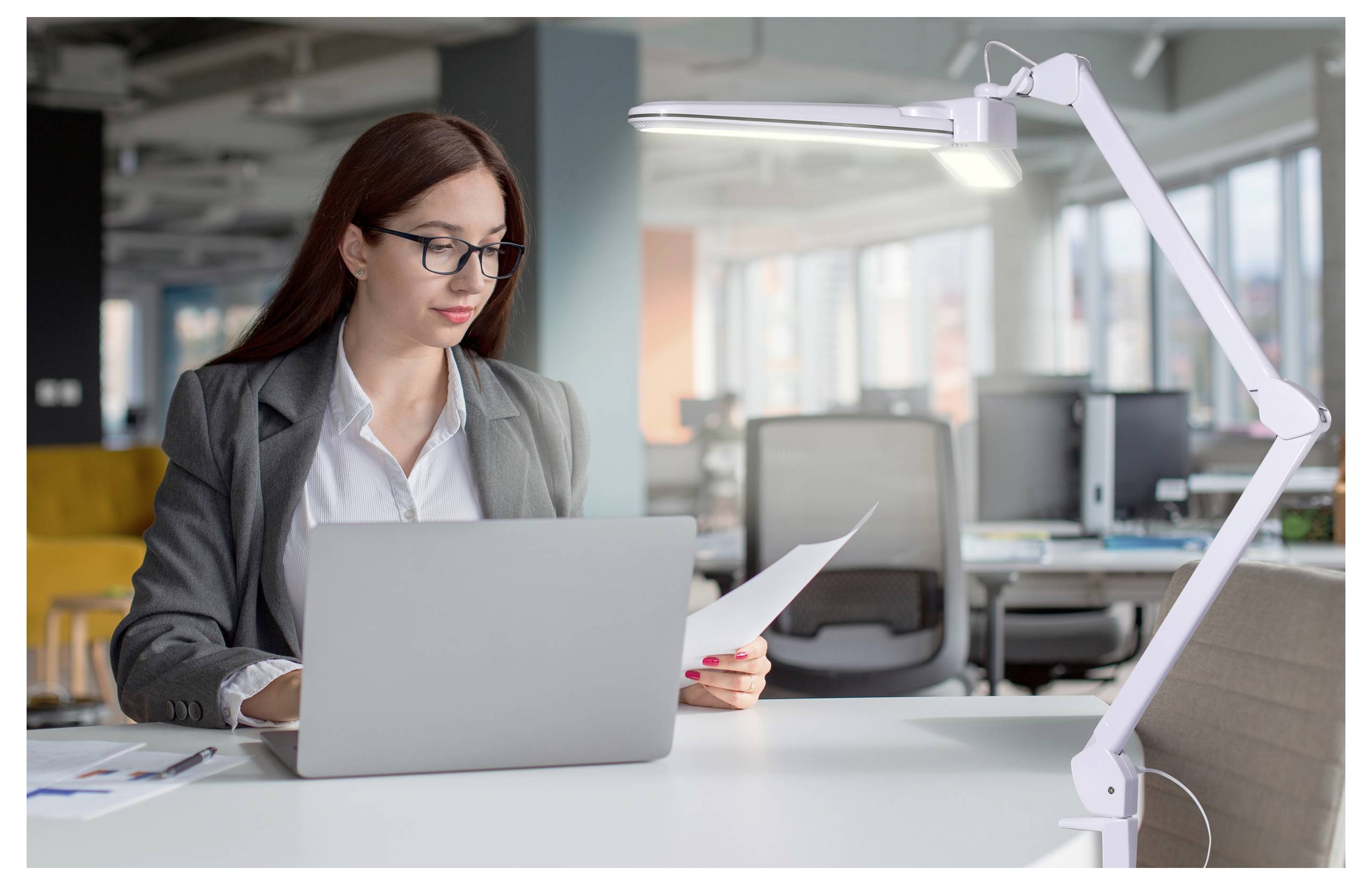 Eine Person in einem grauen Blazer arbeitet an einem Laptop in einer modernen Büroumgebung und schaut auf ein Dokument. Eine helle Schreibtischlampe beleuchtet den Arbeitsbereich.