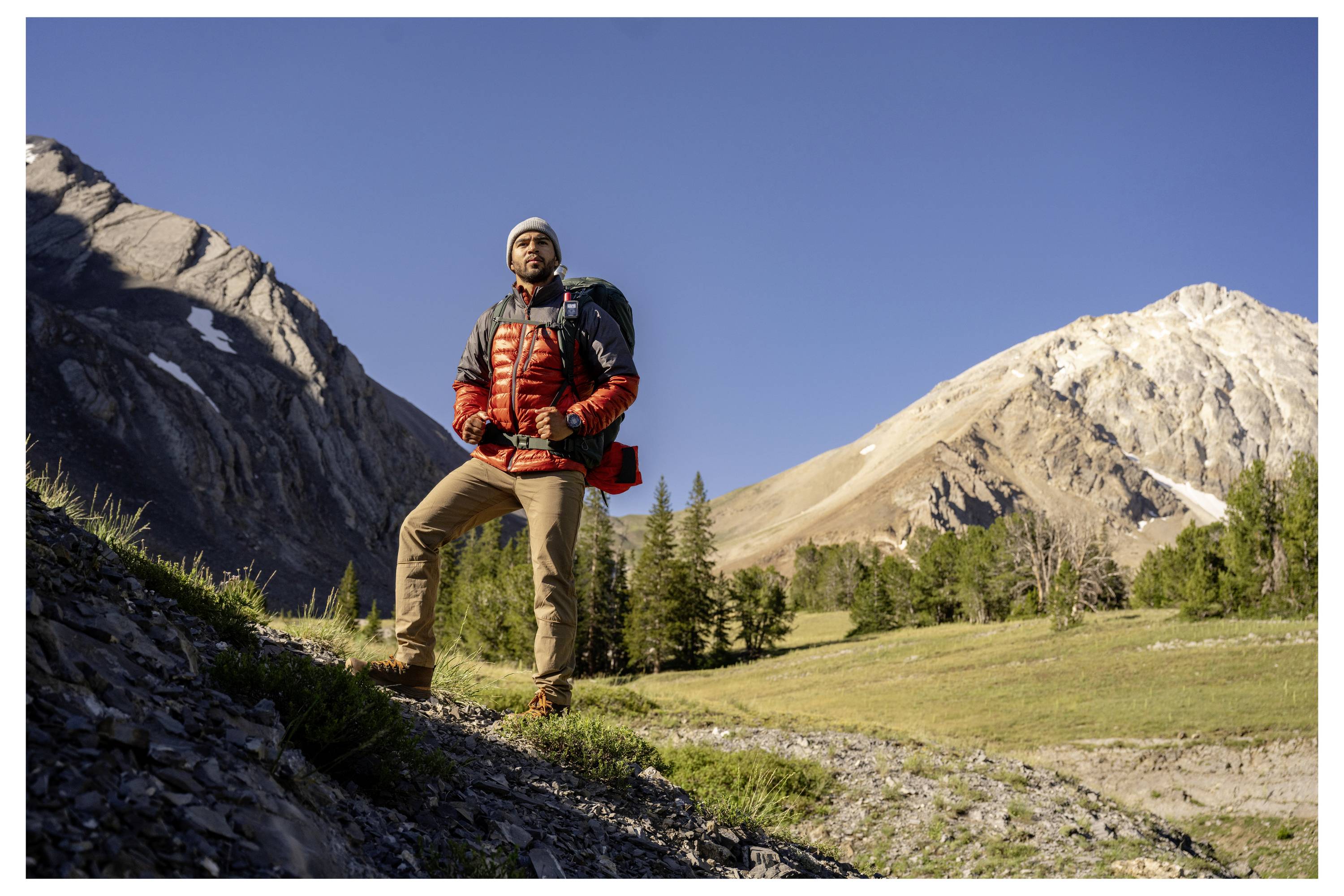 Eine Person in Wanderausrüstung steht selbstbewusst auf einem felsigen Pfad, mit einem klaren blauen Himmel und majestätischen Bergen im Hintergrund.