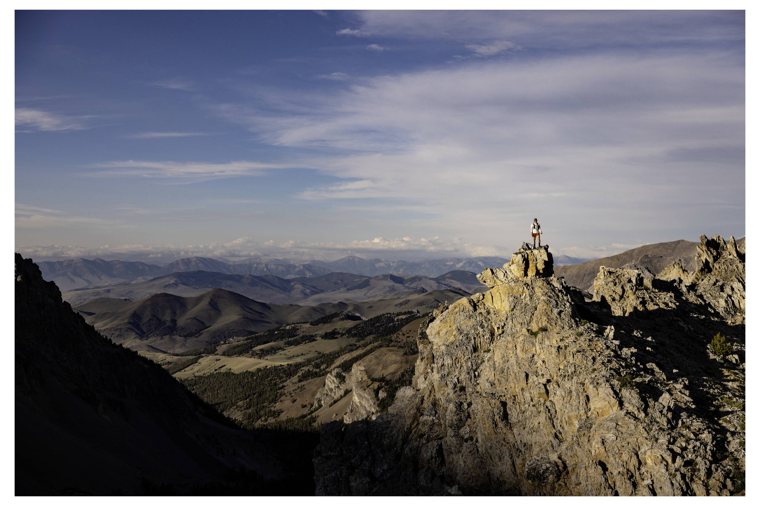 Eine Person steht auf einem felsigen Berggipfel und blickt über eine weite Landschaft von Tälern und fernen Bergen unter einem blauen Himmel mit Wolken.