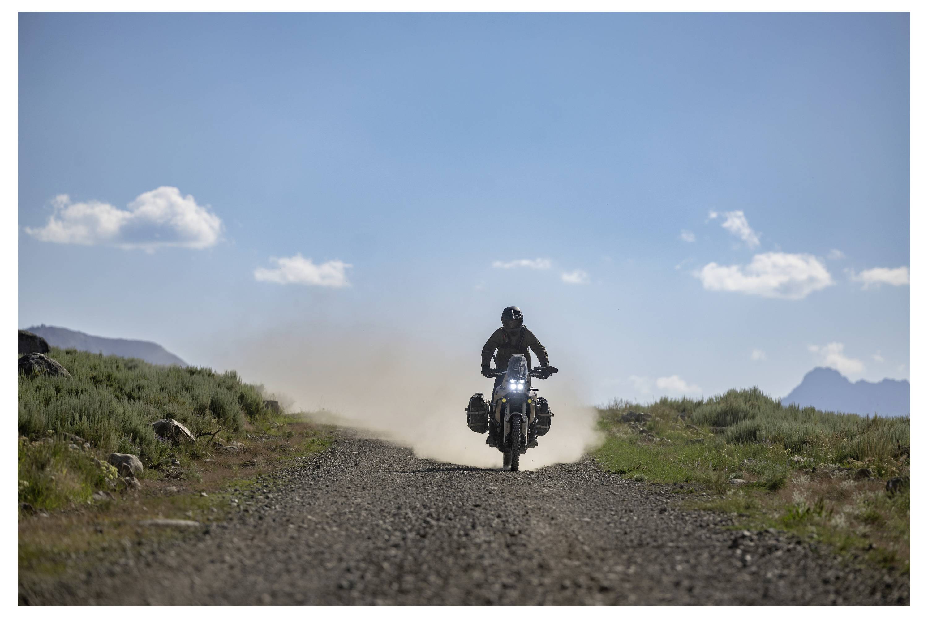 Eine Person fährt Motorrad auf einem Schotterweg, umgeben von offener Landschaft mit Gras und Bergen unter einem klaren blauen Himmel mit Wolken.