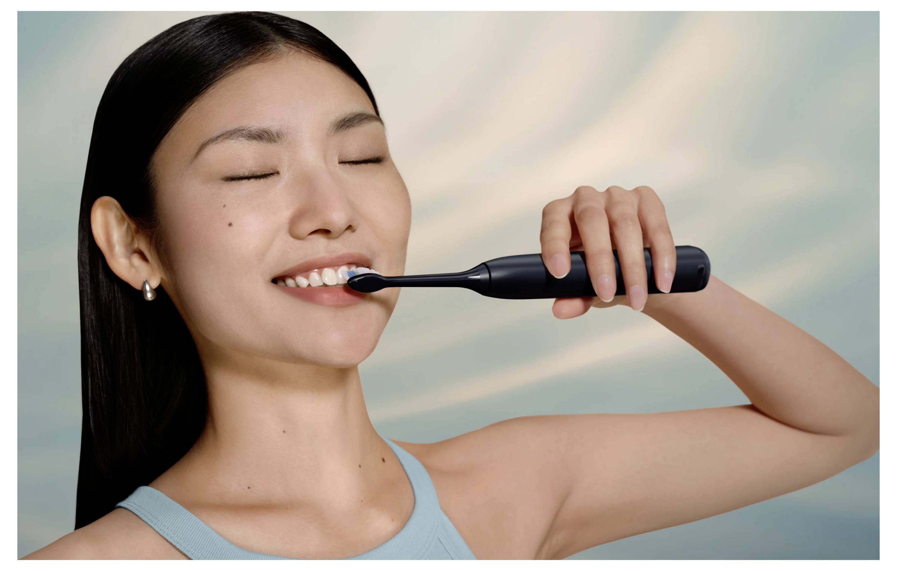 Frau, die sich mit einer elektrischen Zahnbürste die Zähne putzt, lächelt und trägt ein hellblaues Oberteil vor einem weichen Himmelhintergrund.