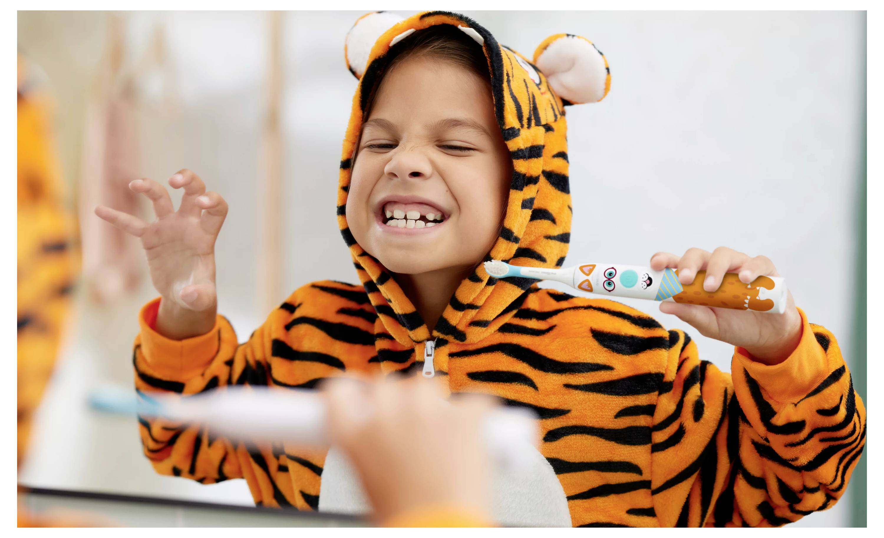 Ein Kind in einem Tigerkostüm putzt sich spielerisch die Zähne und macht eine Krallengeste vor einem Spiegel.