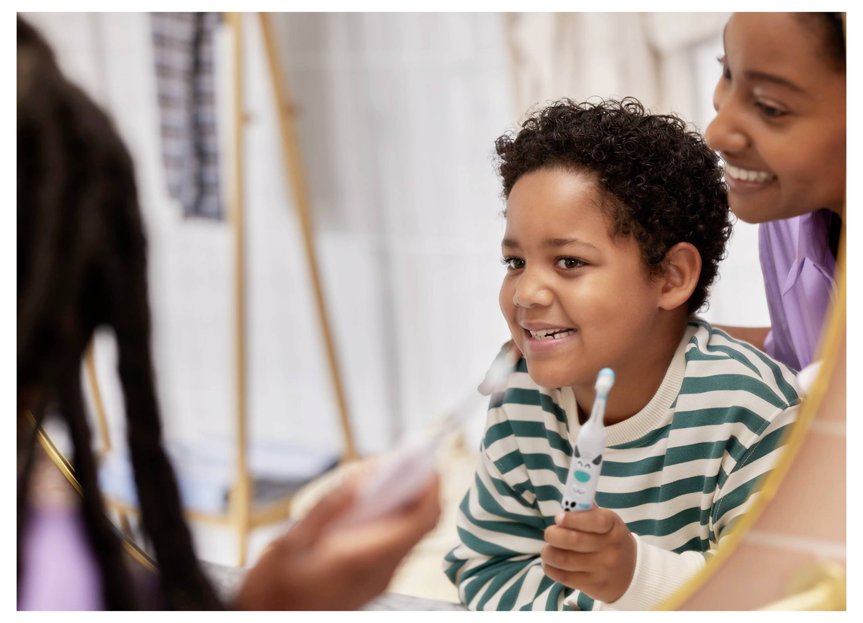 Ein Kind mit lockigem Haar lächelt vor einem Badezimmerspiegel und hält dabei eine Zahnbürste, während ein Erwachsener in der Nähe steht und die Zahnpflege ermutigt.