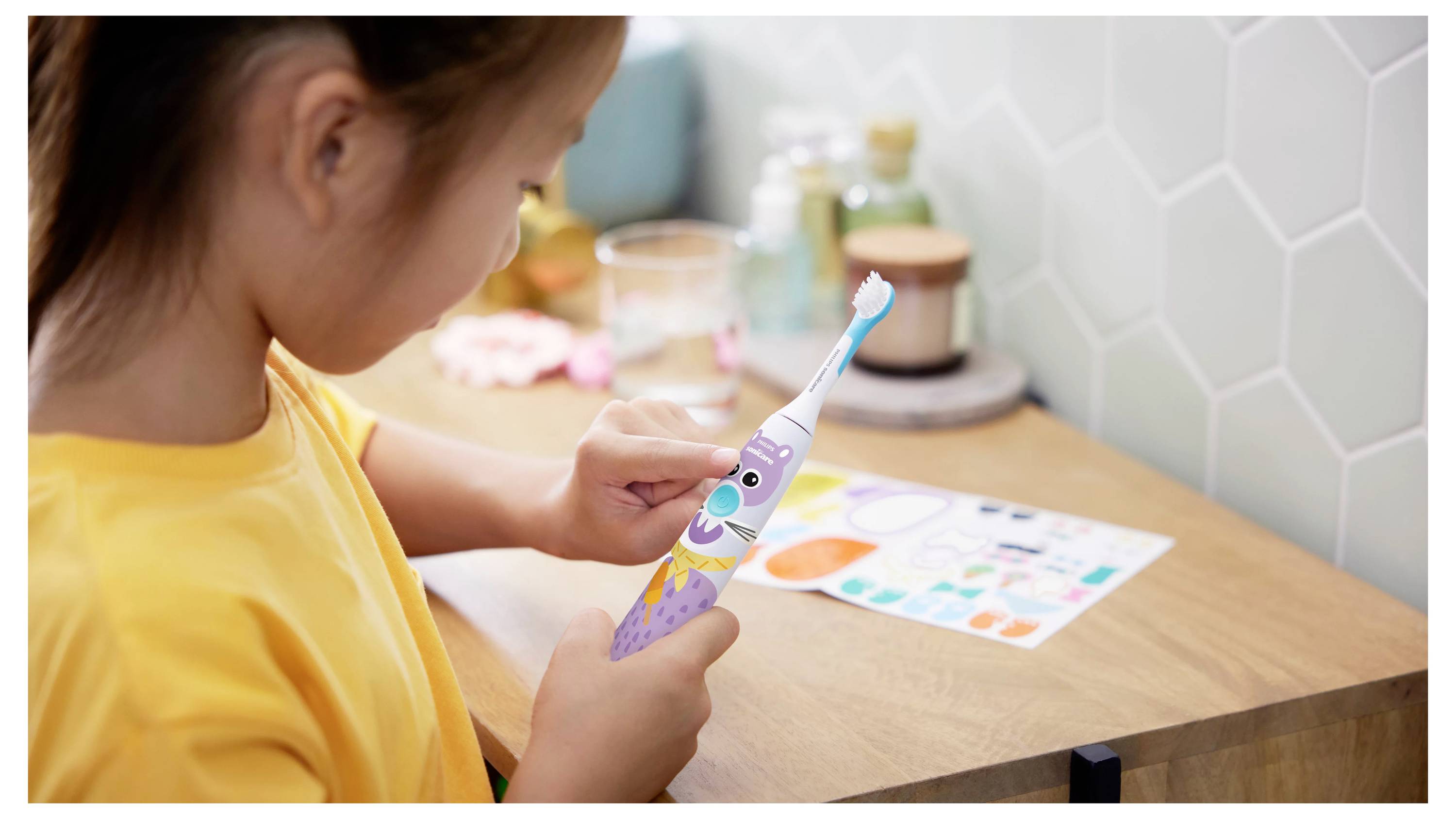 Ein Kind in einem gelben Shirt betrachtet eine bunte elektrische Zahnbürste mit Comicfiguren und steht dabei an einem Badezimmerwaschbecken mit verschiedenen Gegenständen.