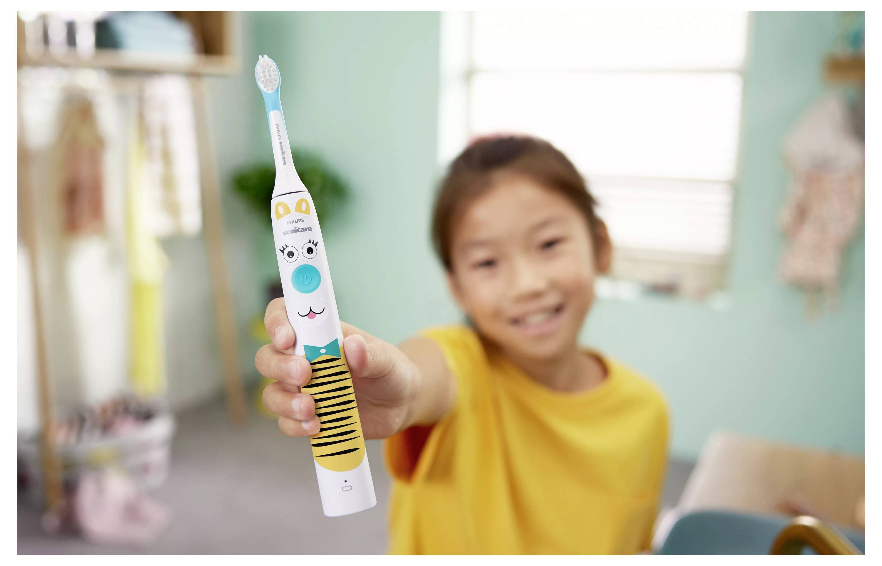 Ein Kind in einem gelben Shirt hält eine bunte Zahnbürste mit Tigerdesign und lächelt in einem sanft beleuchteten Raum.