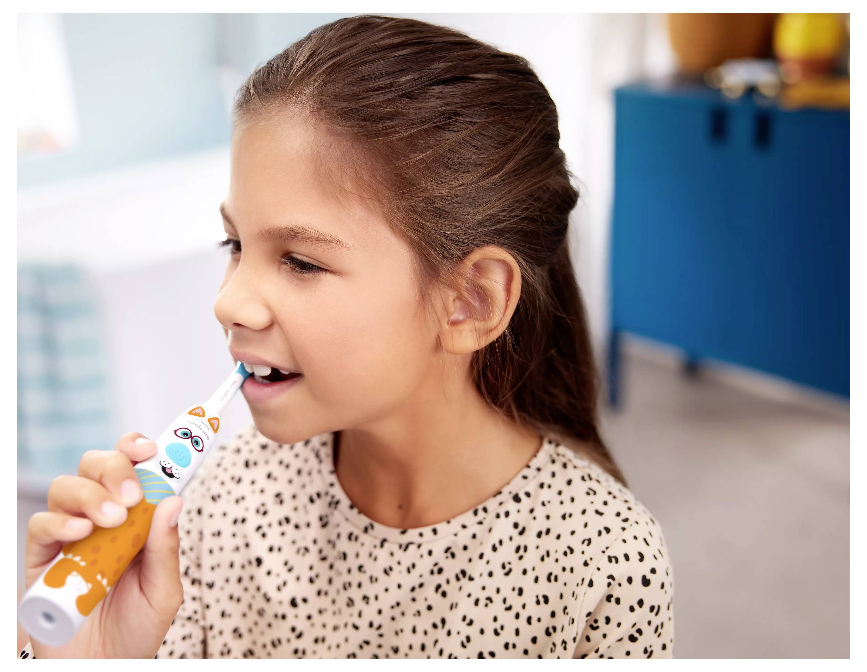 Junges Mädchen putzt Zähne mit einer bunten Zahnbürste, seitlich blickend, trägt ein gemustertes Shirt, im Innenraum.