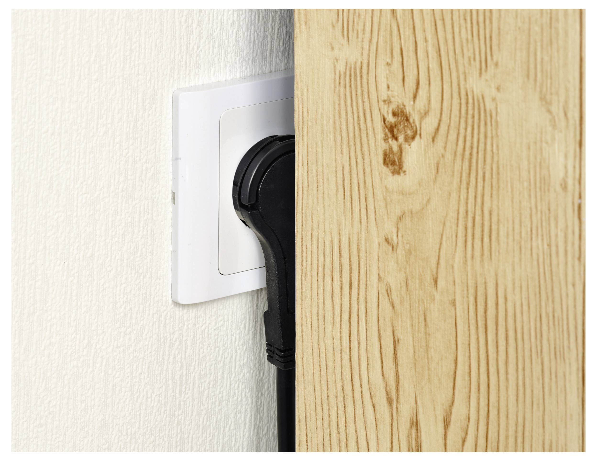 Ein schwarzer Stromstecker wird in eine weiße Steckdose eingefügt, wobei sich eine Holzverkleidung in unmittelbarer Nähe befindet und wenig Platz für den Stecker vorhanden ist.
