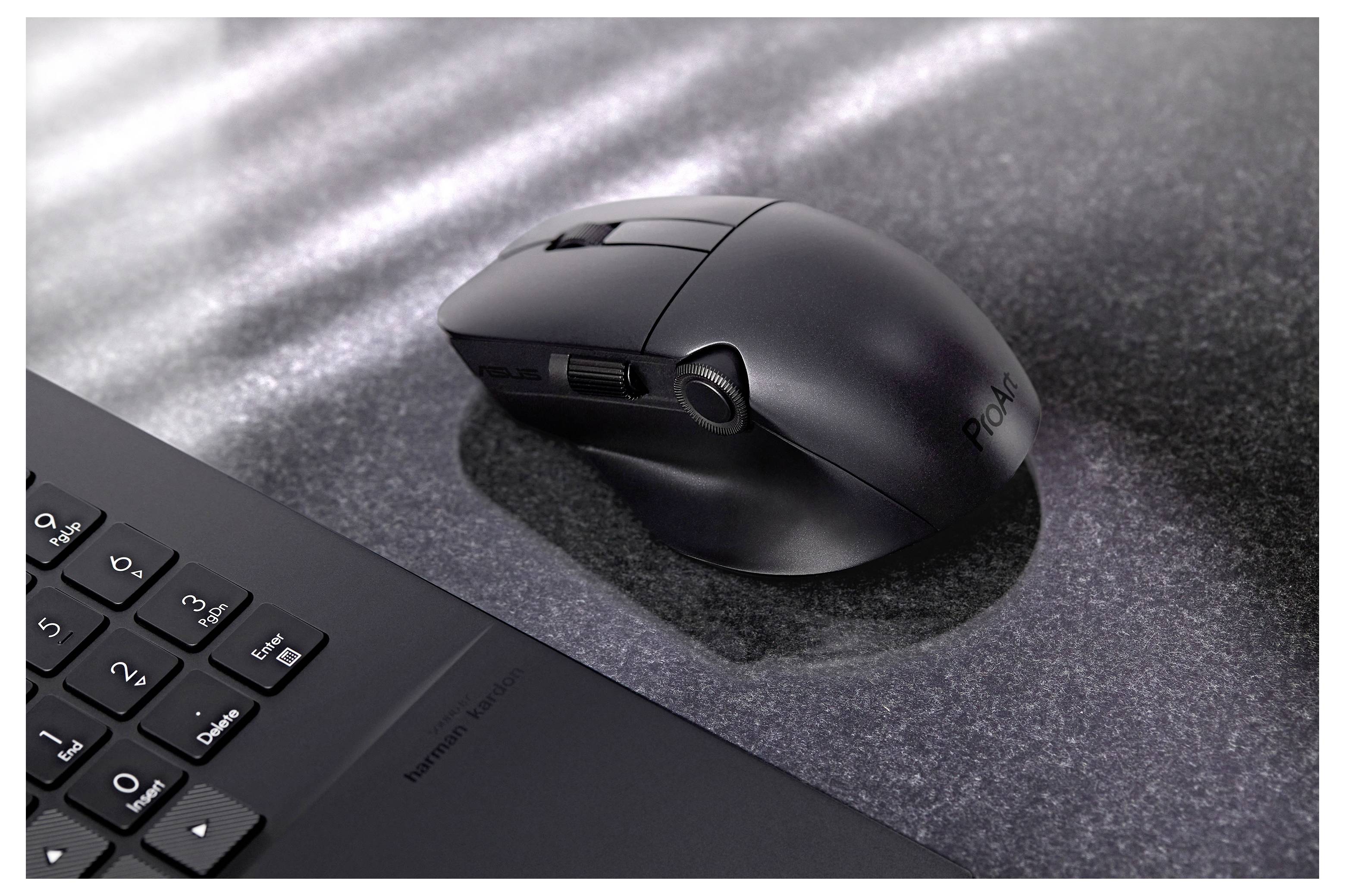 Eine schwarze kabellose Maus auf einer dunklen strukturierten Oberfläche, neben einer teilweisen Ansicht einer Laptop-Tastatur. Das Licht fällt quer über die Oberfläche.