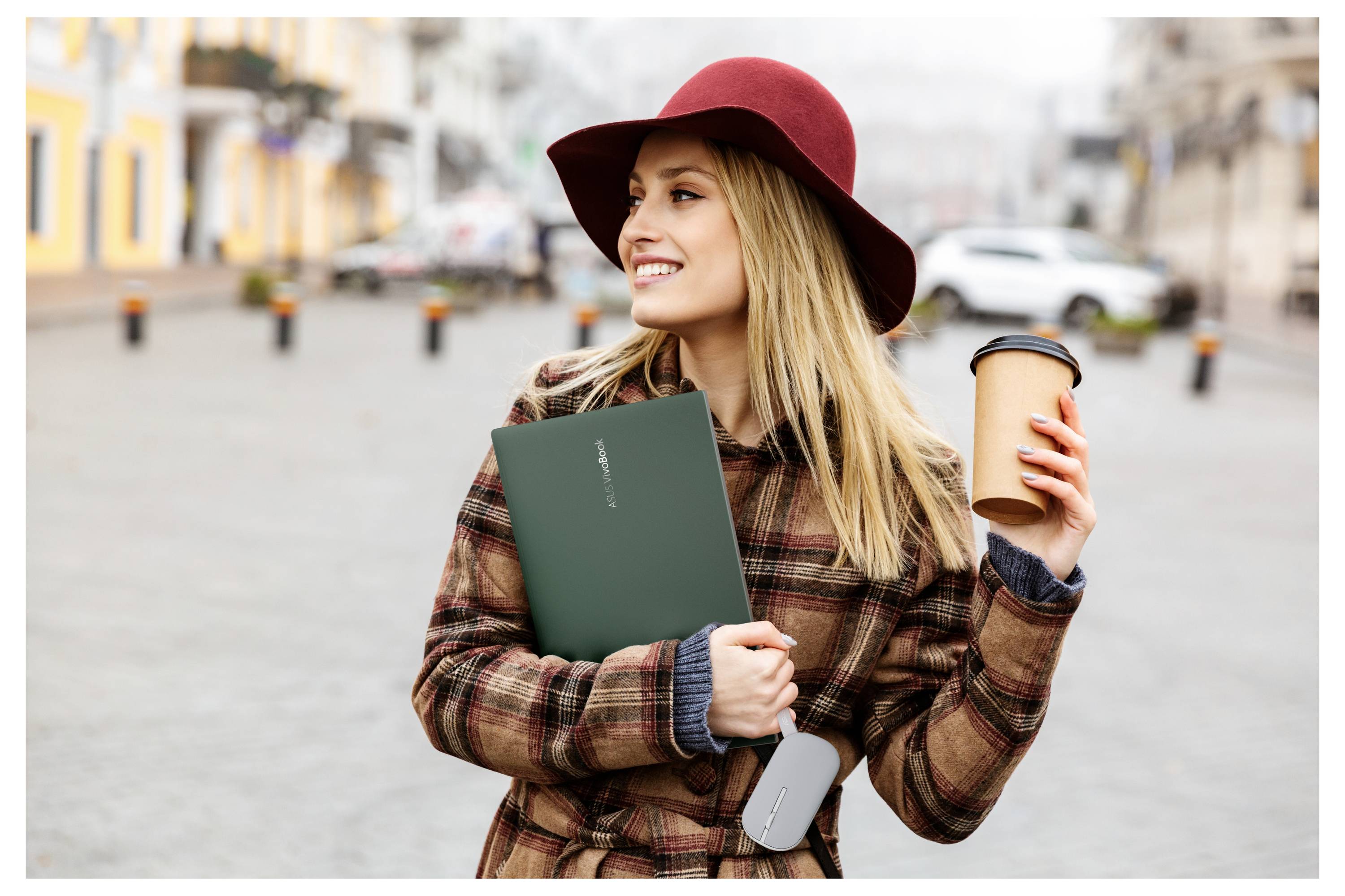 Eine Frau in einem Karomantel und rotem Hut steht im Freien, hält eine Kaffeetasse und einen grünen Ordner, lächelt, mit unscharfem städtischem Hintergrund.