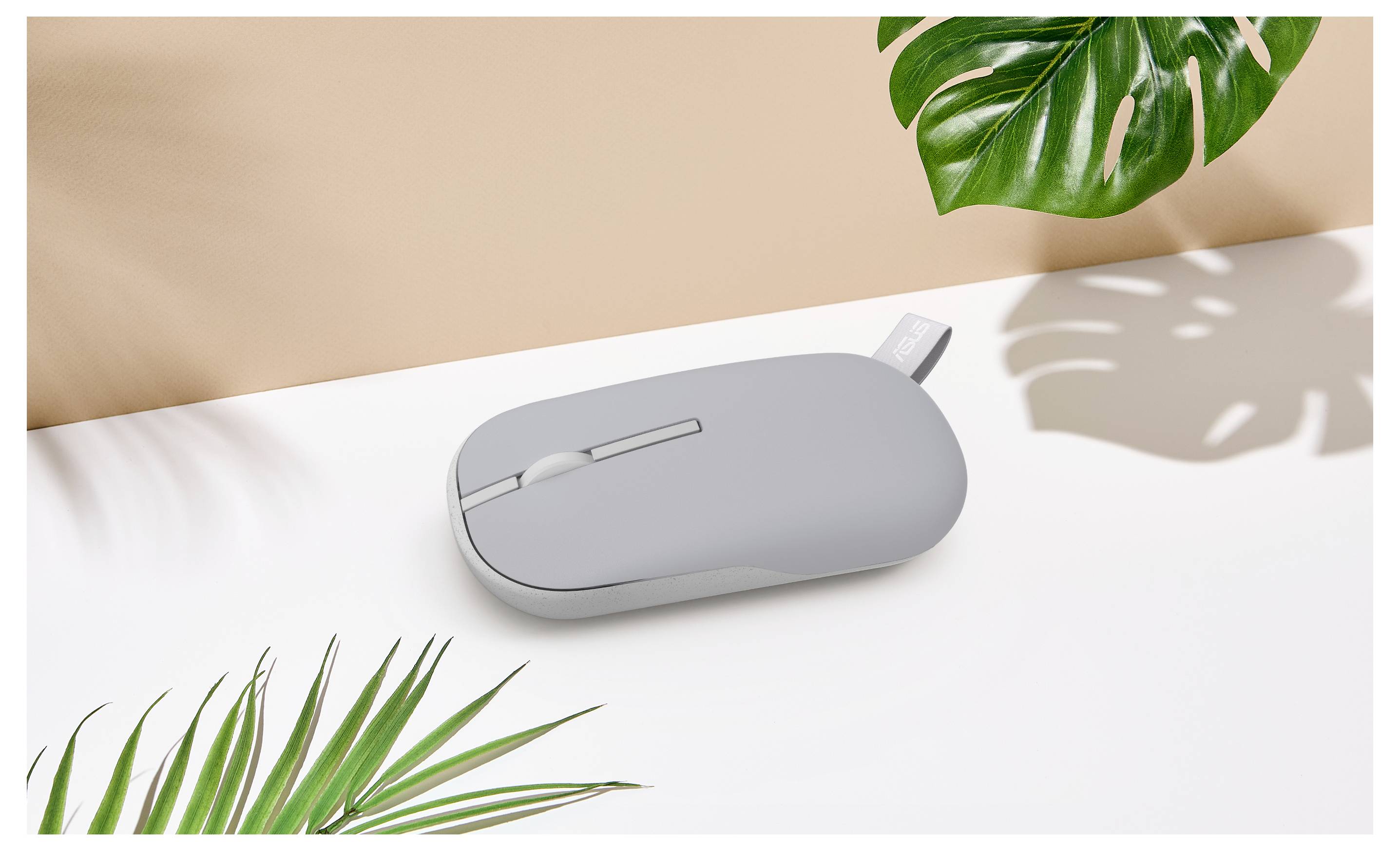 Eine minimalistische graue Computermaus auf einer weißen Oberfläche, umgeben von grünen tropischen Blättern, die weiche Schatten werfen.