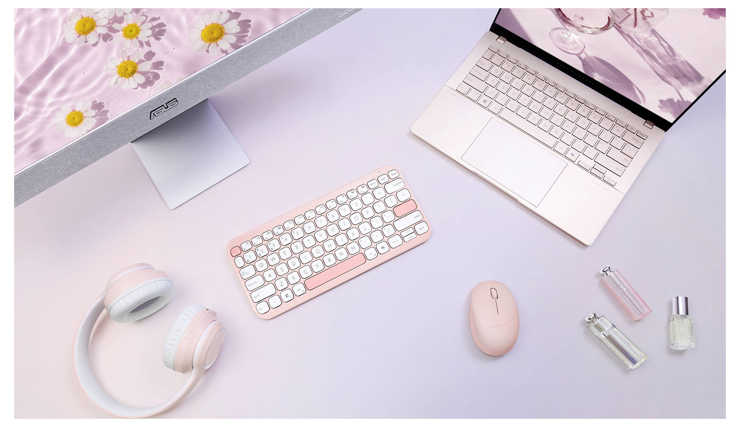 Ein pastellfarbener Arbeitsplatz mit einem rosa Laptop, einer drahtlosen Tastatur und Maus, Kopfhörern und Parfümflakons auf einer hellrosa Oberfläche.