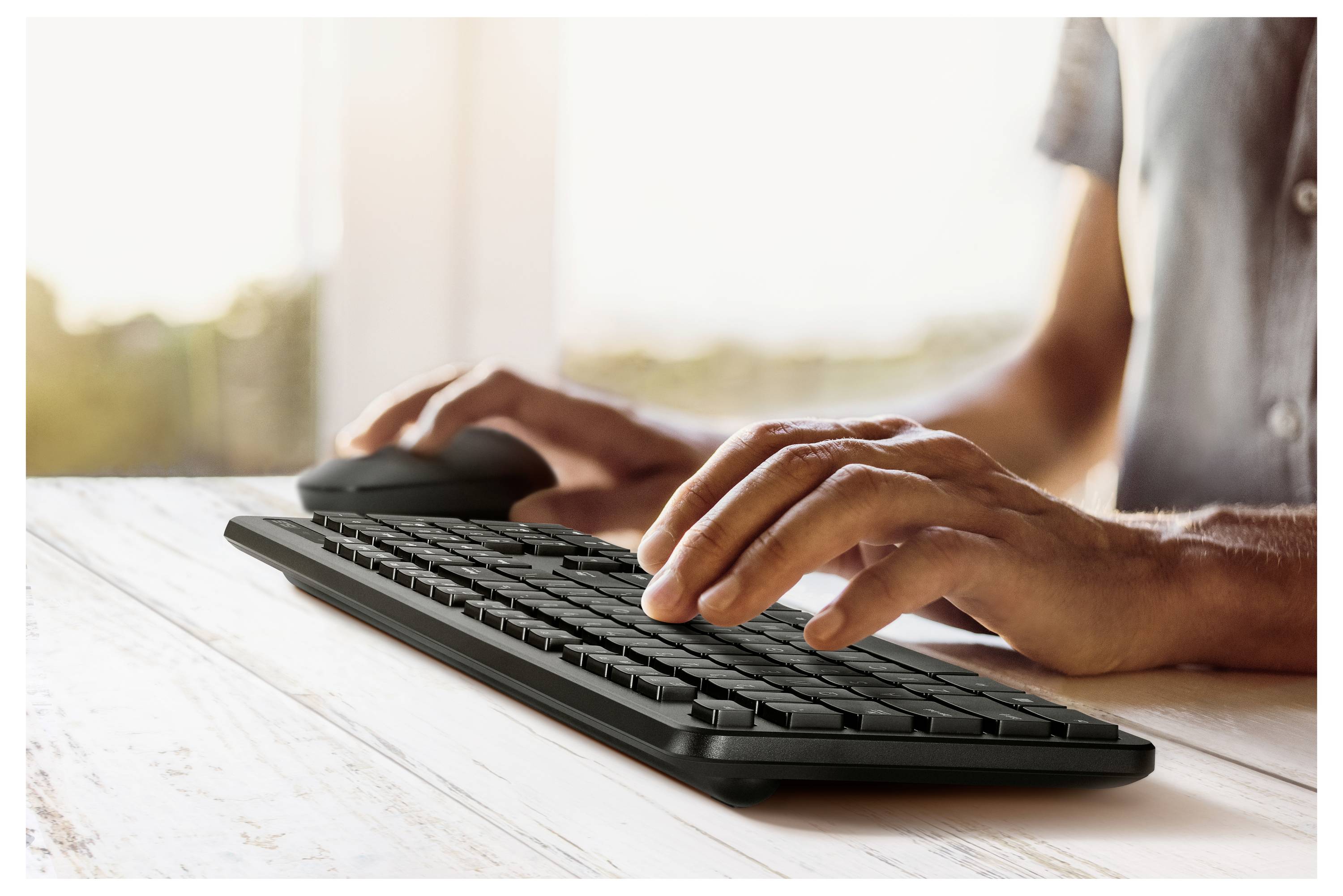 Eine Person, die auf einer drahtlosen Tastatur tippt, mit einer Maus in der Nähe, in einem hellen Raum mit Fensterblick.