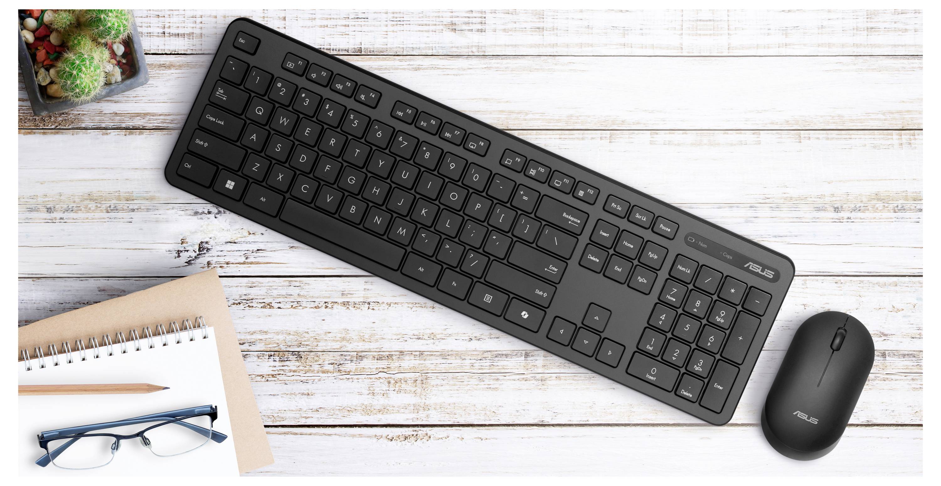 Eine schwarze Tastatur und eine schwarze Maus, die auf einem weißen Holztisch platziert sind, neben einem Notizbuch, einem Stift, einer Brille und kleinen Pflanzen.