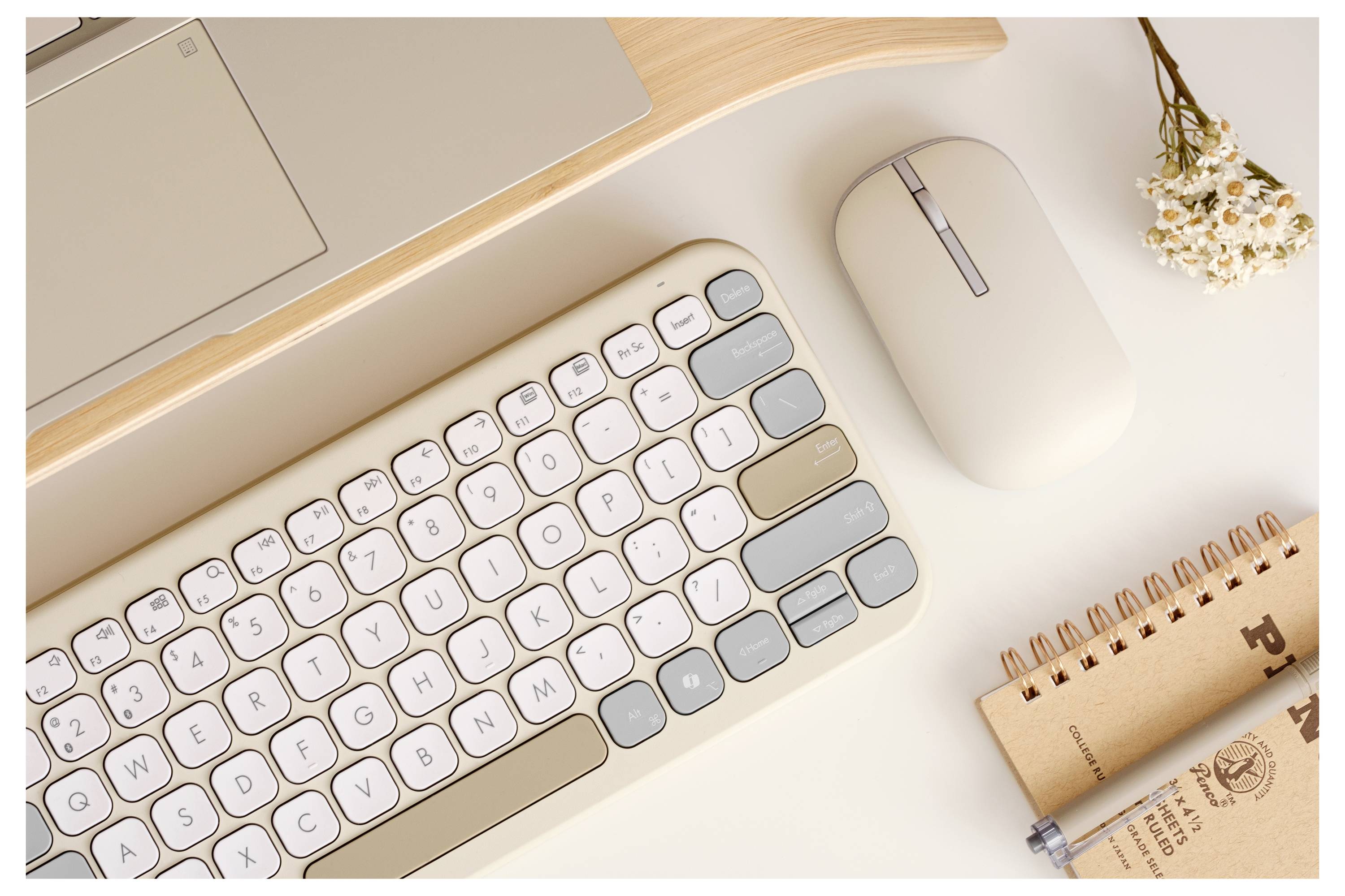 Eine beige Tastatur und Maus neben einem Laptop, mit einem kleinen Blumenstrauß und einem Spiralblock in der Nähe auf einem hellen Schreibtisch.