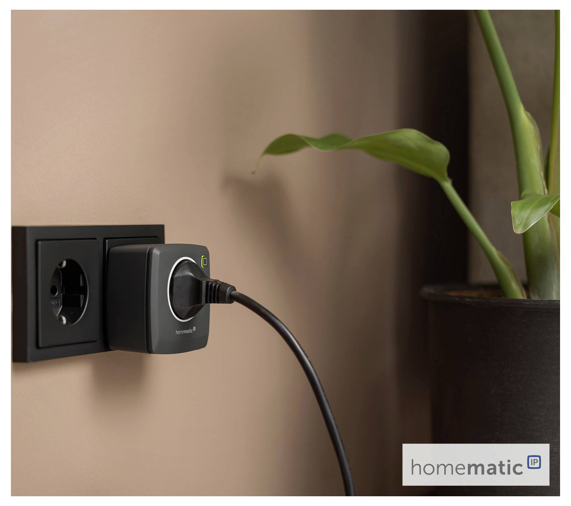 Ein intelligenter Stecker ist mit einer Wandsteckdose neben einer Pflanze verbunden und zeigt ein grünes Licht. Das Logo von 'homematic id' ist in der Ecke sichtbar.