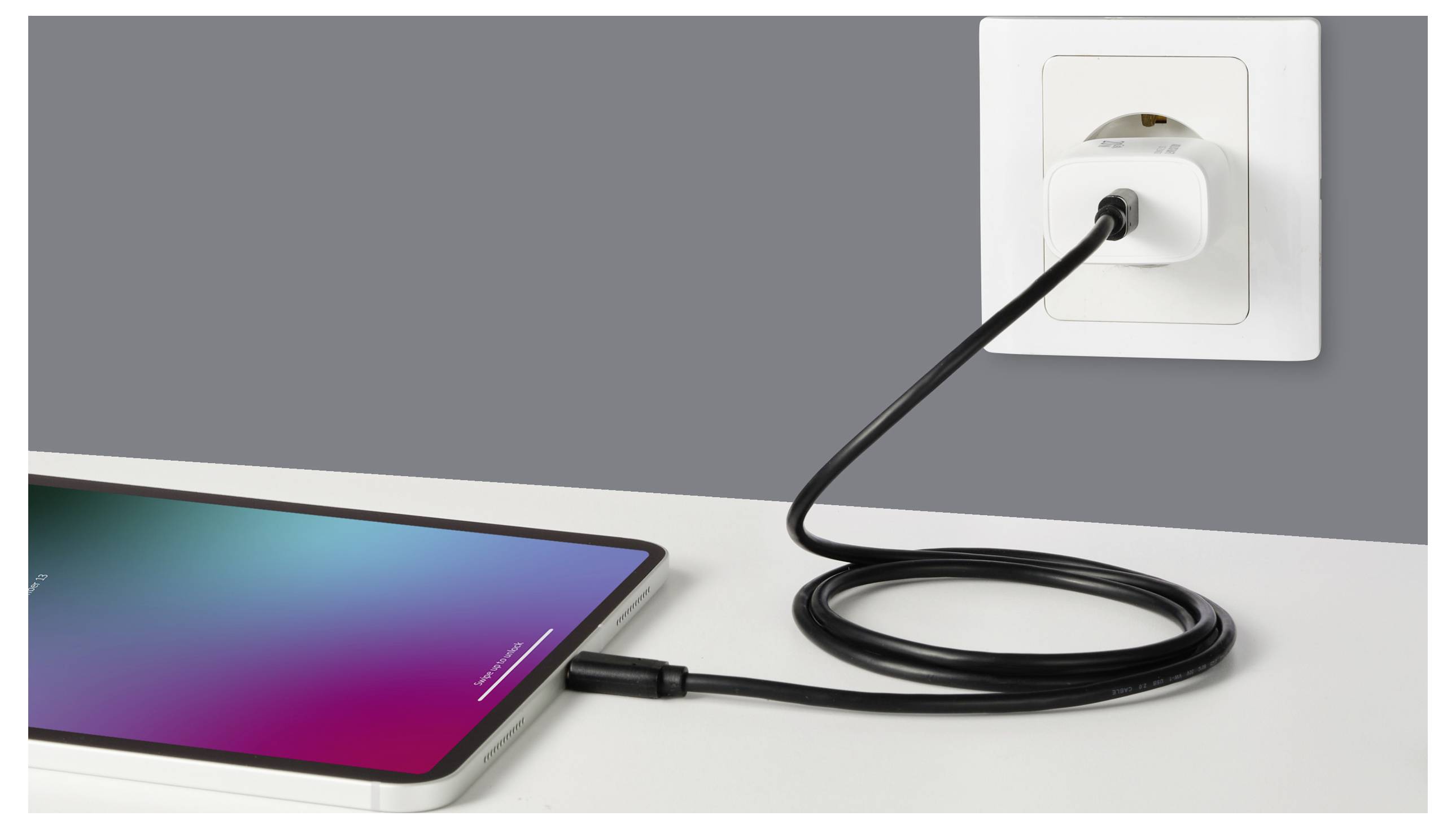 Ein Tablet auf einem Tisch wird aufgeladen; ein schwarzes Kabel verbindet sich mit einem Wandsteckeradapter.