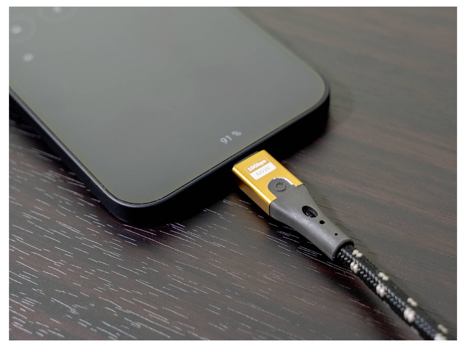 Ein Smartphone wird mit einem geflochtenen Kabel aufgeladen und zeigt auf einer dunklen Holzoberfläche einen Batteriestand von 91% an.