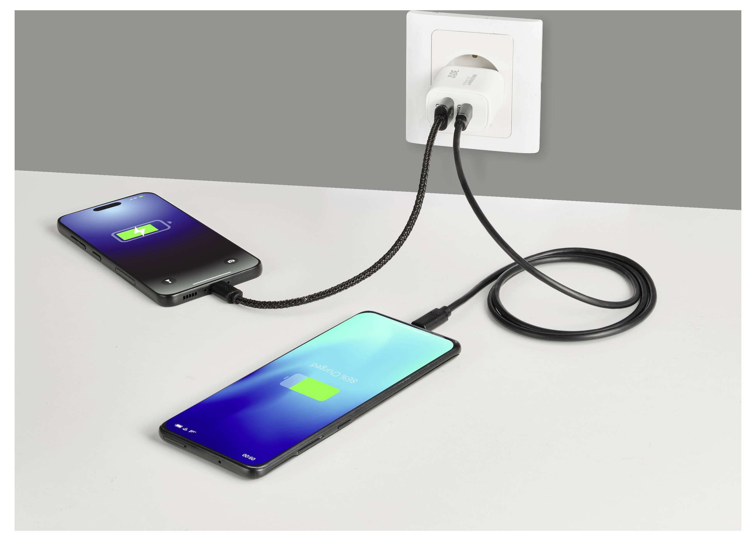 Zwei Smartphones laden über eine Wandsteckdose mit Doppelausgang. Beide Bildschirme zeigen Akku-Ladesymbole an, die aktiven Stromtransfer anzeigen.