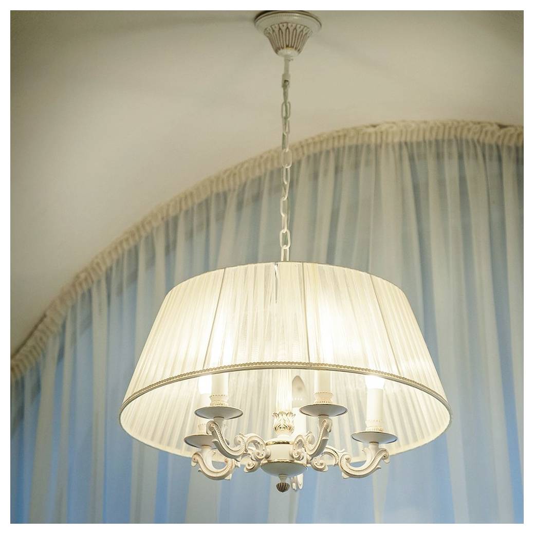 Ein dekorativer Kronleuchter mit fünf kerzenähnlichen Glühbirnen und einem transparenten Lampenschirm, der vor einem Hintergrund aus weichen blauen Vorhängen hängt.