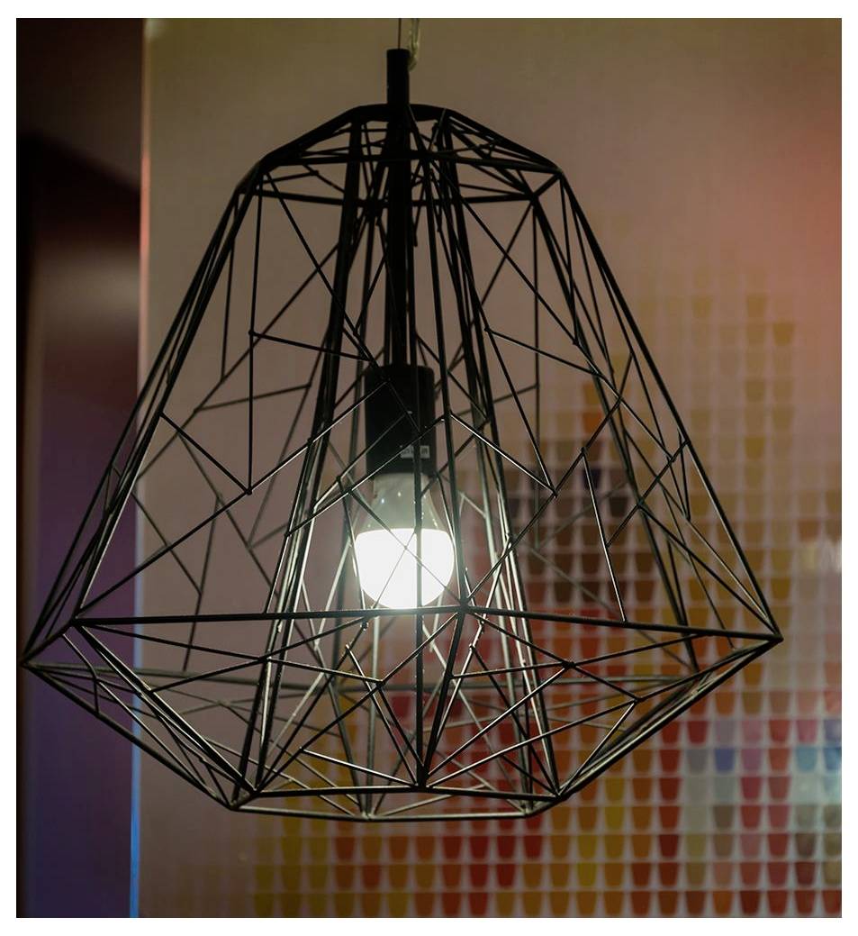 Eine moderne geometrische Drahtgitter-Pendelleuchte mit einer eingeschalteten Glühbirne im Inneren hängt vor einem unscharfen Hintergrund mit einem bunten Mosaikmuster.