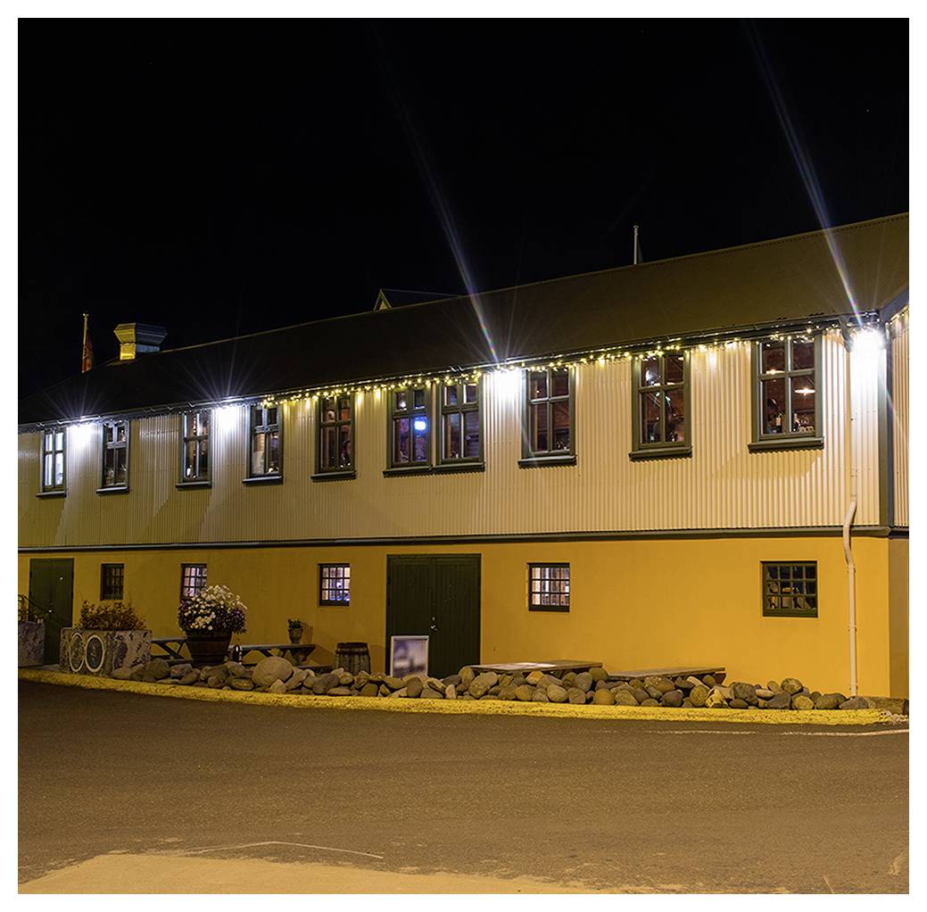 Ein gelb-weißes Gebäude bei Nacht, geschmückt mit Lichterketten und umgeben von einer Steinmauer, das eine warme, einladende Atmosphäre schafft.