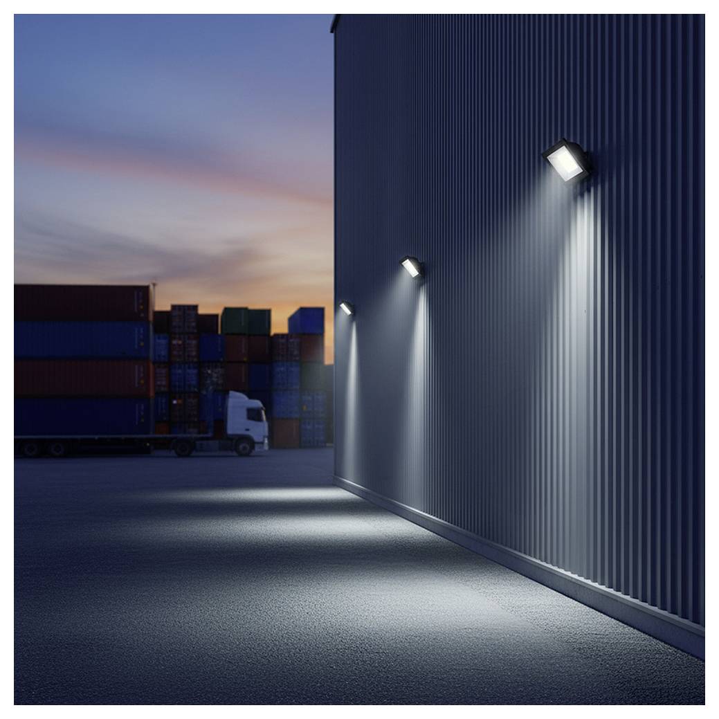 Ein schwach beleuchtetes Industriegebiet in der Dämmerung mit einer Gebäudewand, die von drei Lichtern beleuchtet wird. Gestapelte Versandcontainer und ein Lastkraftwagen sind sichtbar.