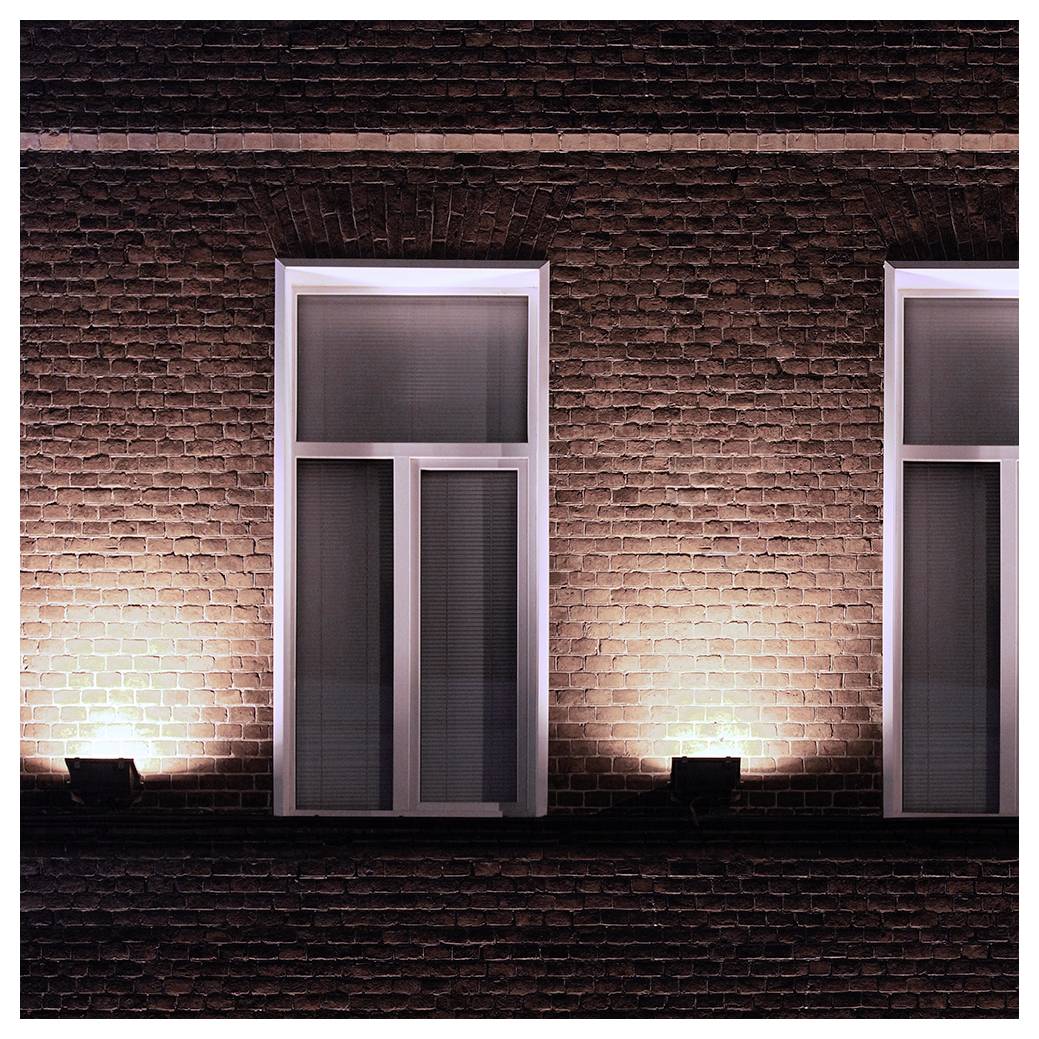 Zwei beleuchtete Fenster an einer Ziegelwand, jedes mit geschlossenen Jalousien. Warmes Licht wirft Schatten und hebt die Textur der Ziegelsteine hervor.