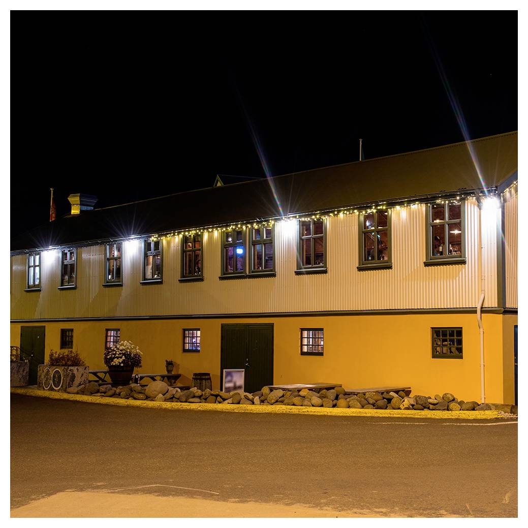 Ein warm beleuchtetes Gebäude mit Lichterketten bei Nacht. Große Fenster offenbaren ein gemütliches Inneres. Ein mit Steinen gesäumter Weg verläuft entlang der Fassade.