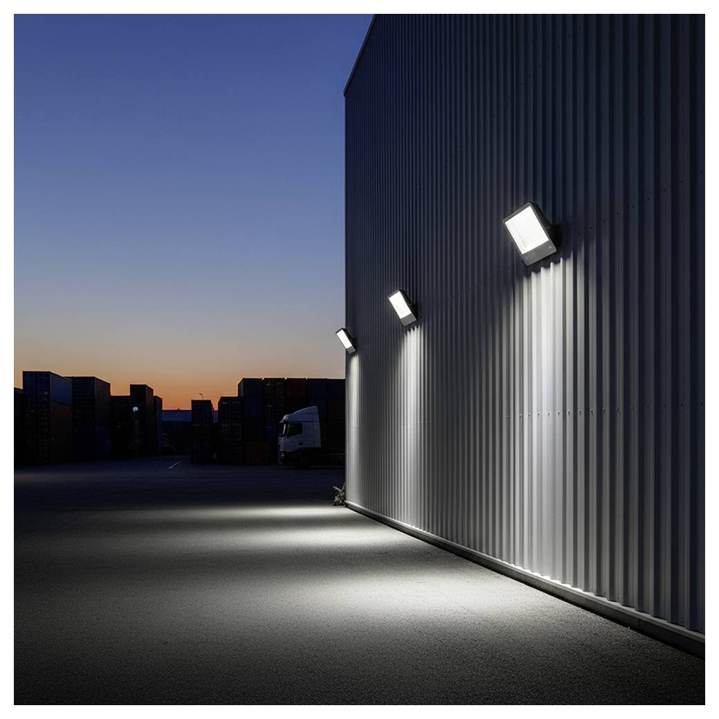 Außenleuchten beleuchten ein Wellblechgebäude in der Dämmerung und werfen Schatten auf den Boden, mit Schiffscontainern in der Ferne.