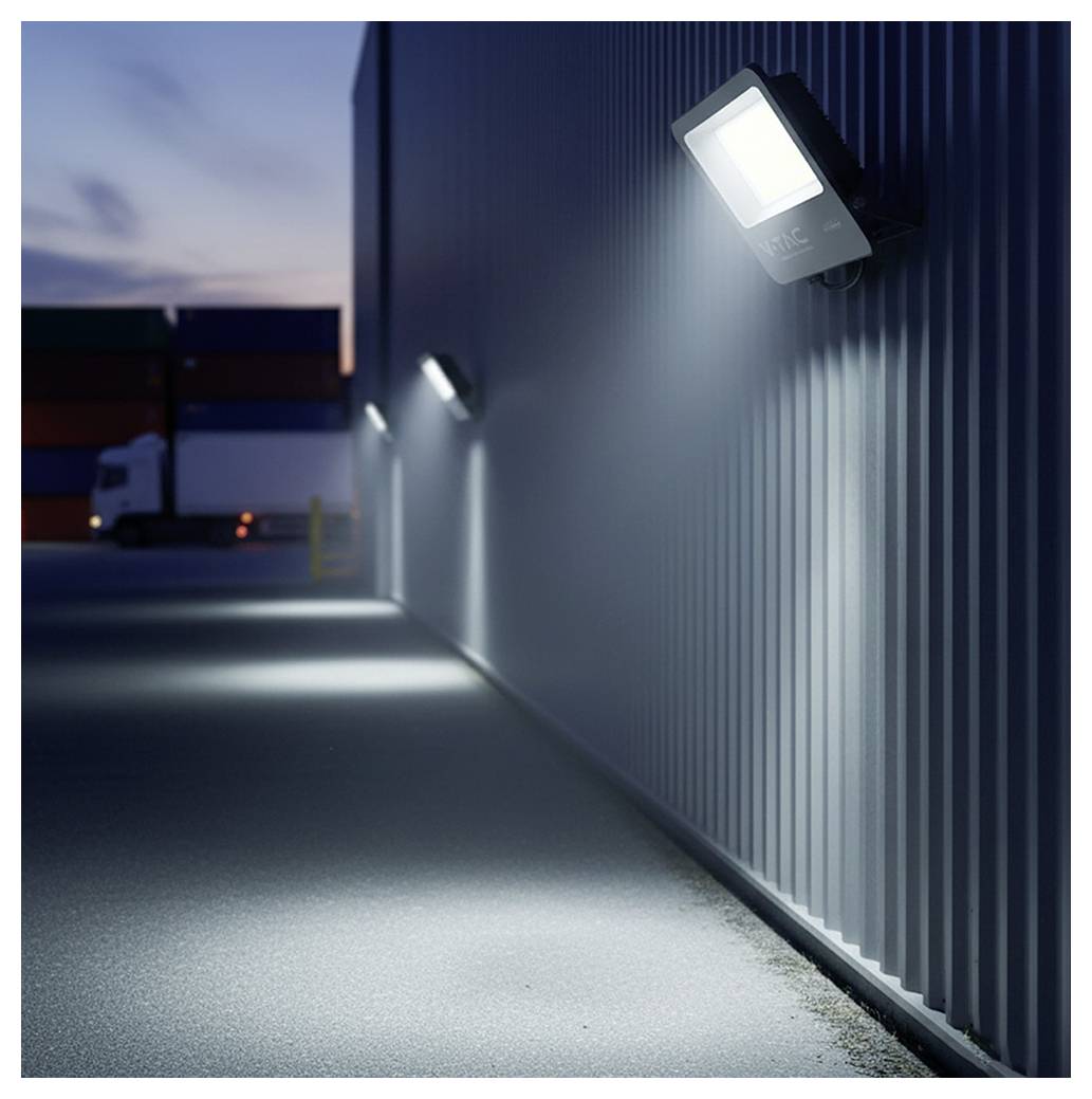 Parkplatz in der Abenddämmerung mit hellen, an der Wand montierten Leuchten, die den Bereich beleuchten. Versandcontainer und ein Lastkraftwagen sind in der Ferne sichtbar.