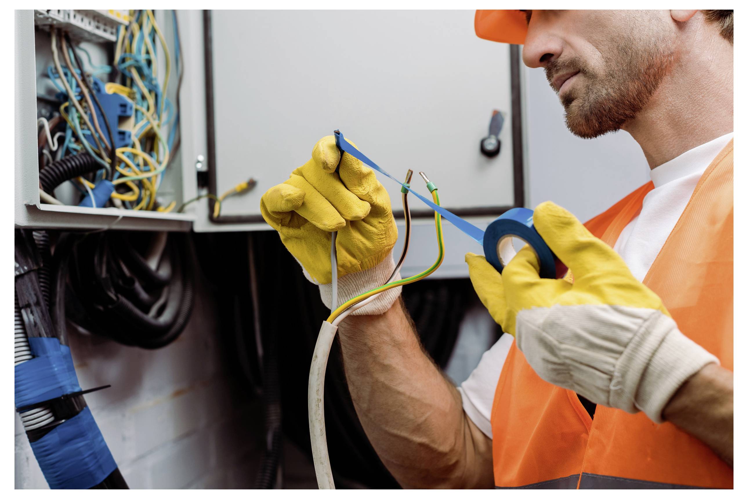 Ein Techniker in Schutzausrüstung verwendet blaues Elektroband und repariert Drähte in einem Schaltschrank.