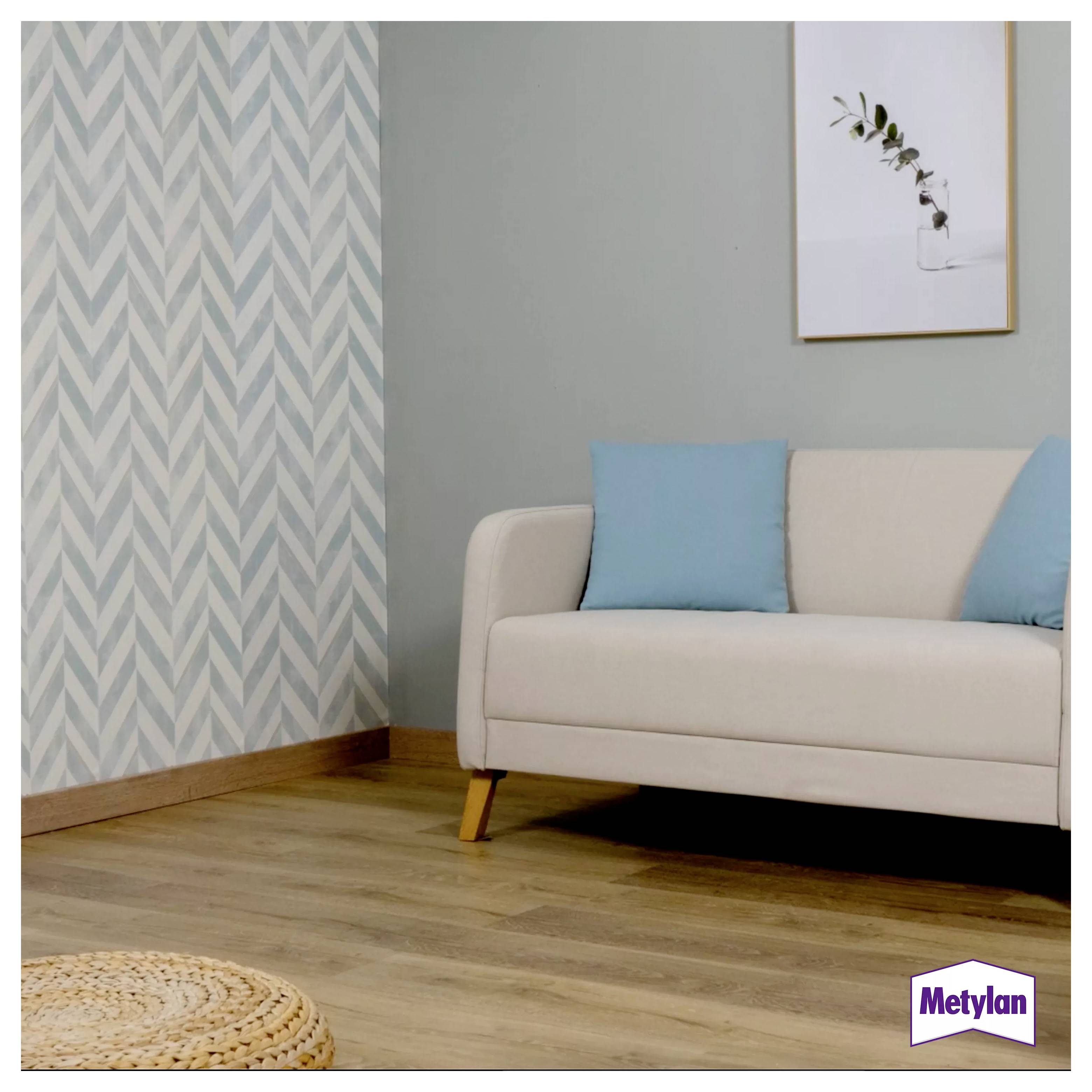 Eine gemütliche Ecke im Wohnzimmer mit einem hellen Sofa und blauen Kissen, Holzboden, zickzackmusterter Tapete, einem gerahmten Blattdruck und einem gewebten Teppich.