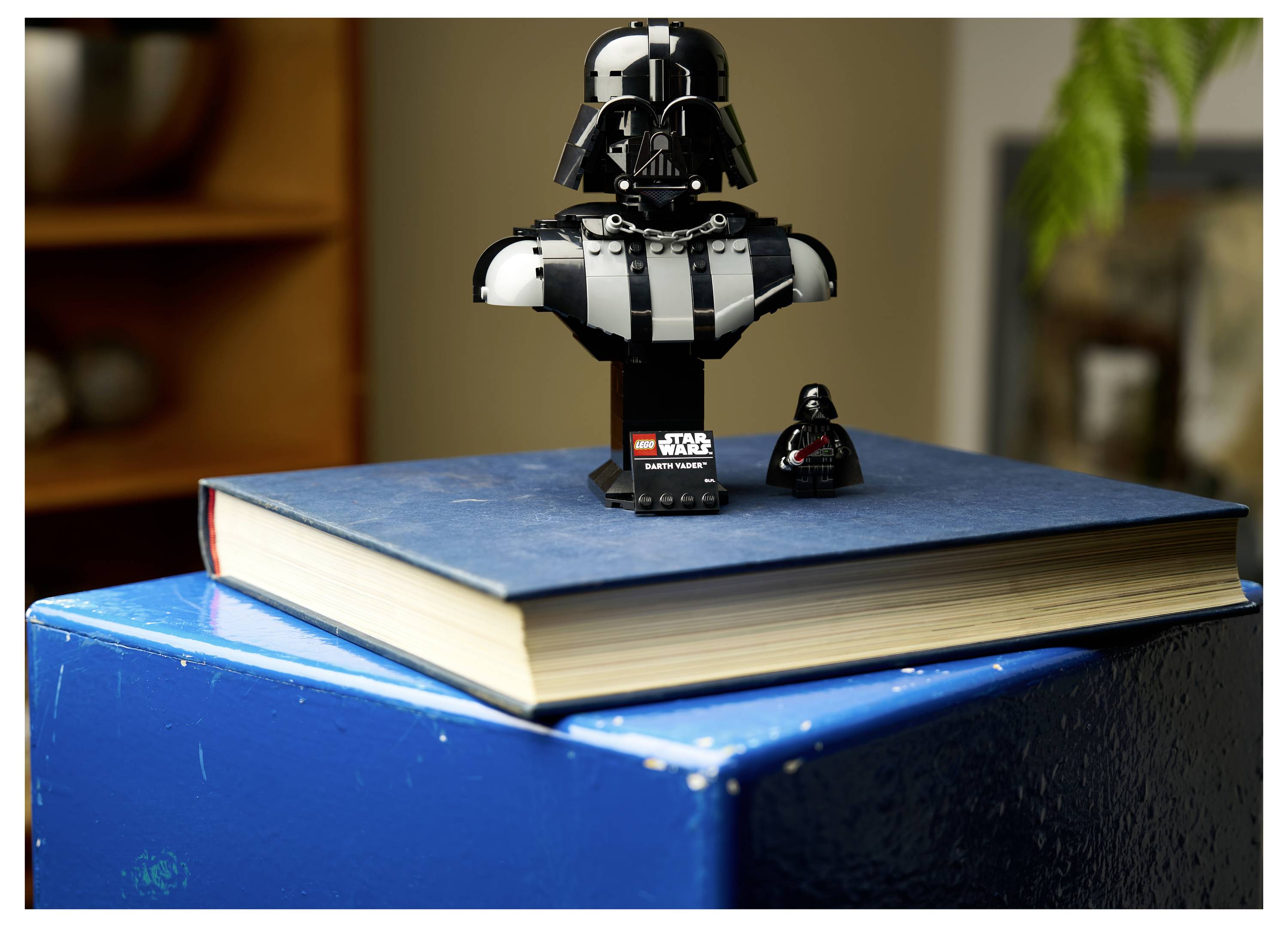 Eine LEGO-Büste einer dunkelhelmierten Figur und eine kleinere Figur ruhen auf einem großen geschlossenen Buch mit blauem Umschlag.