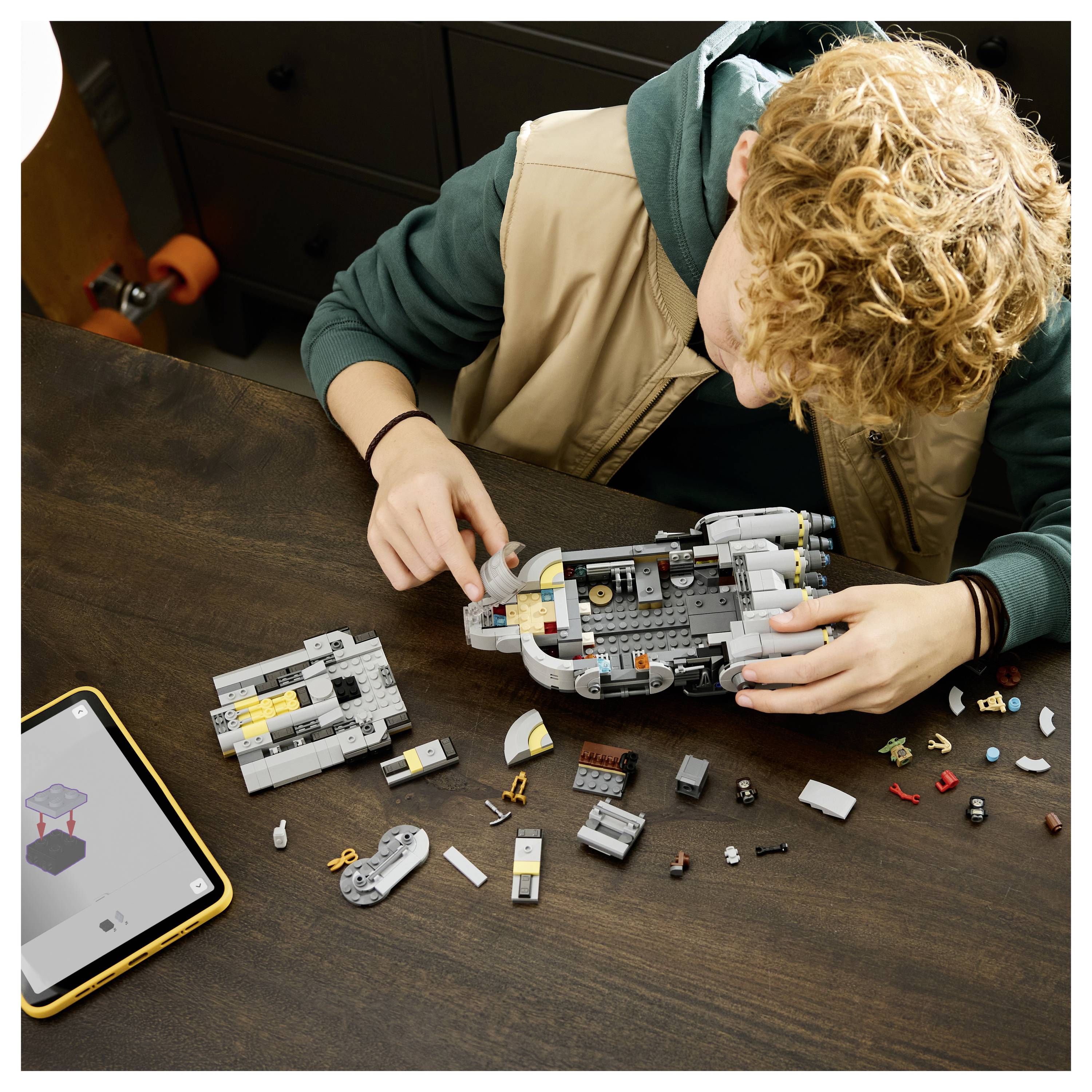Eine Person baut ein Raumschiffmodell aus verstreuten Kunststoffbausteinen auf einem Tisch auf, während ein Tablet eine Schritt-für-Schritt-Anleitung anzeigt.