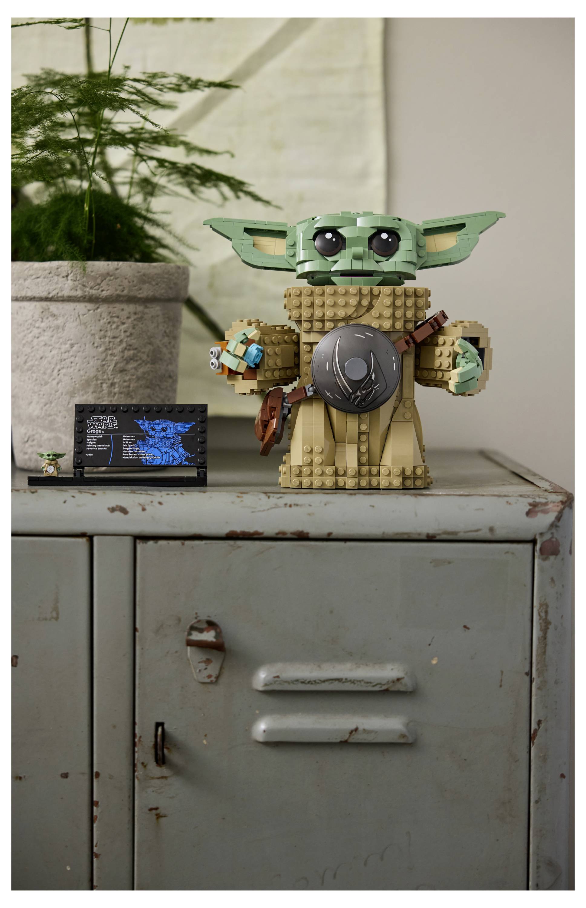 Eine Lego-Figur, die einer kleinen grünen Außerirdischen mit großen Ohren ähnelt, steht auf einem Metallschrank neben einer Topfpflanze. Eine kleine Anzeigetafel steht daneben.