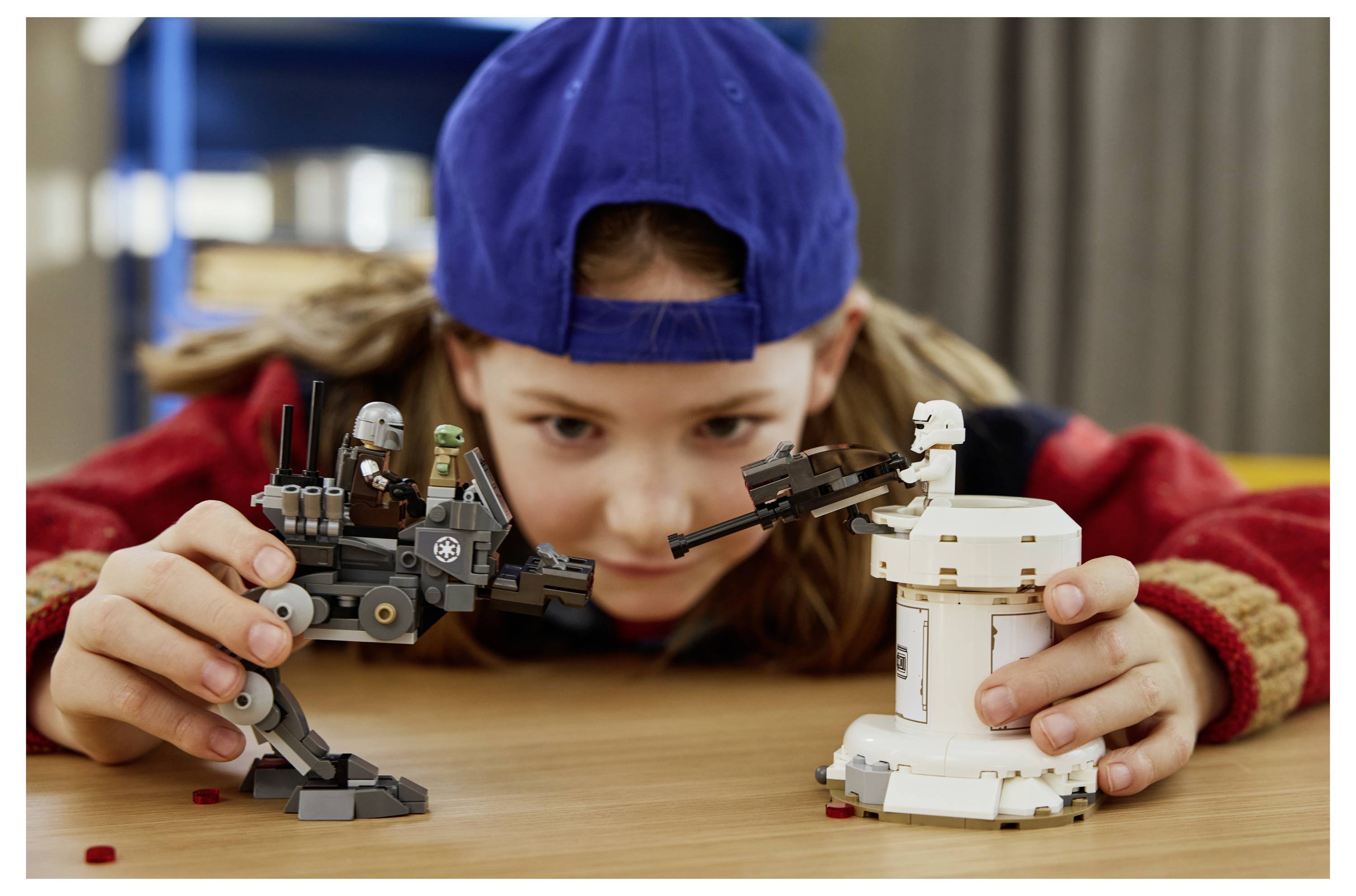 Ein Kind mit einer blauen Mütze und einer roten Jacke spielt mit zwei Star Wars-LEGO-Sets, einem grauen Walker und einem kleinen weißen Geschütz, auf einem Tisch.